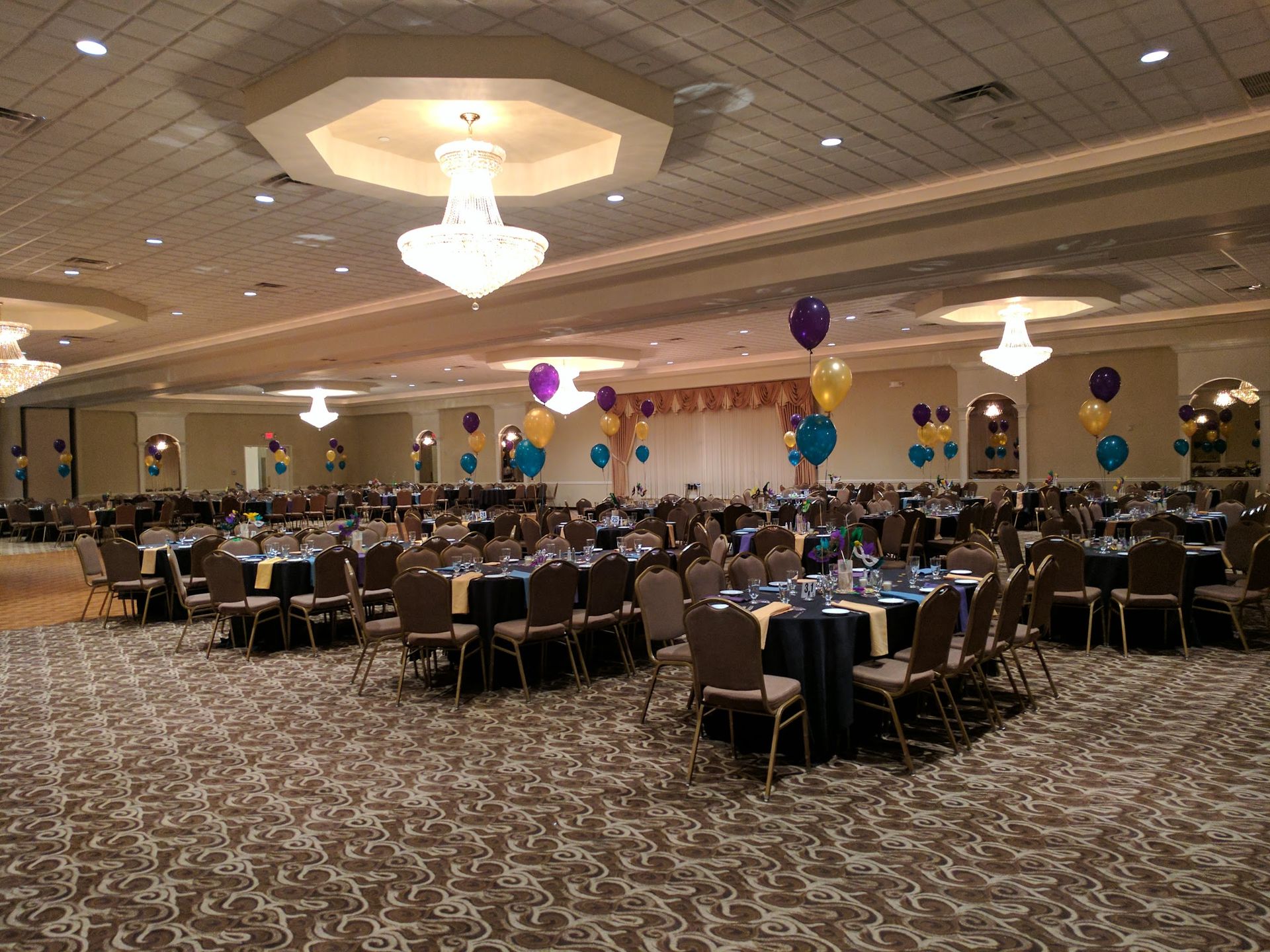 Ballroom set for a banquet with round tables, black chairs, and colorful balloons under chandeliers.