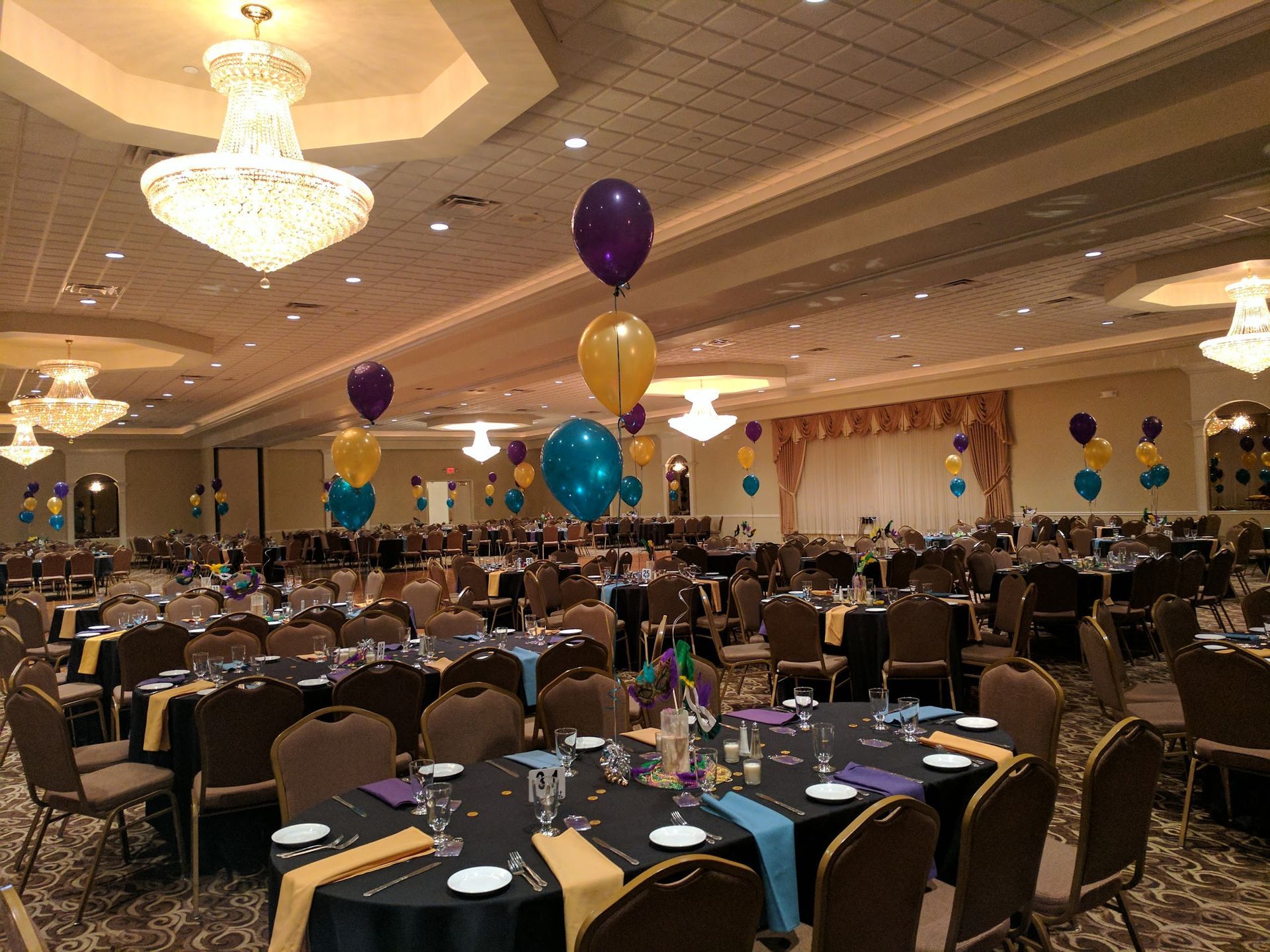 Banquet hall with round tables, chairs, chandeliers, and colorful balloons set for an event