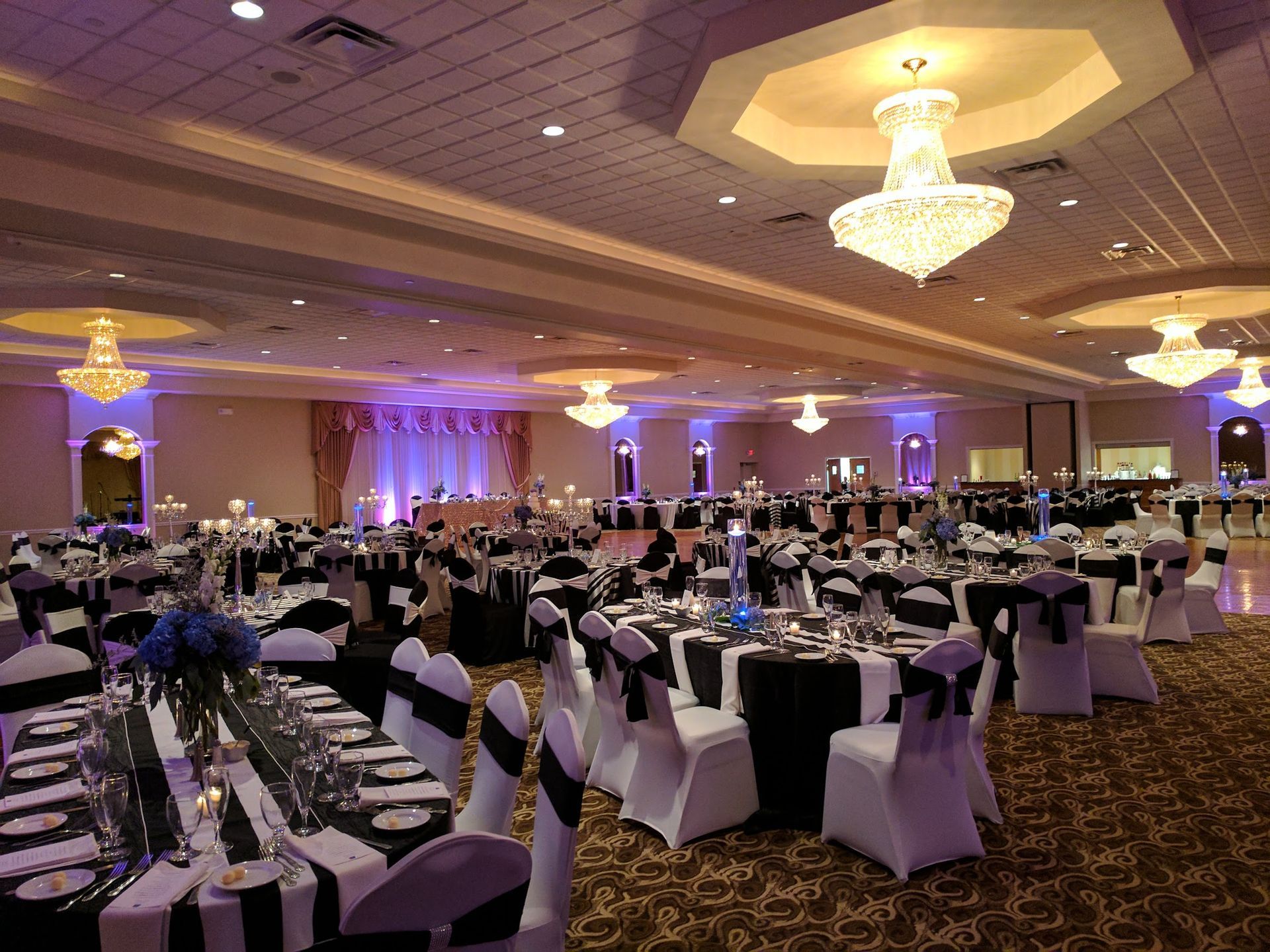Elegant banquet hall set with round tables, black-and-white chairs, and chandeliers under purple lighting