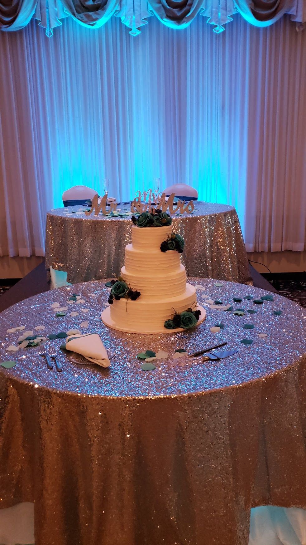 Wedding cake on a sequined table with blue-lit draped backdrop and floral centerpiece