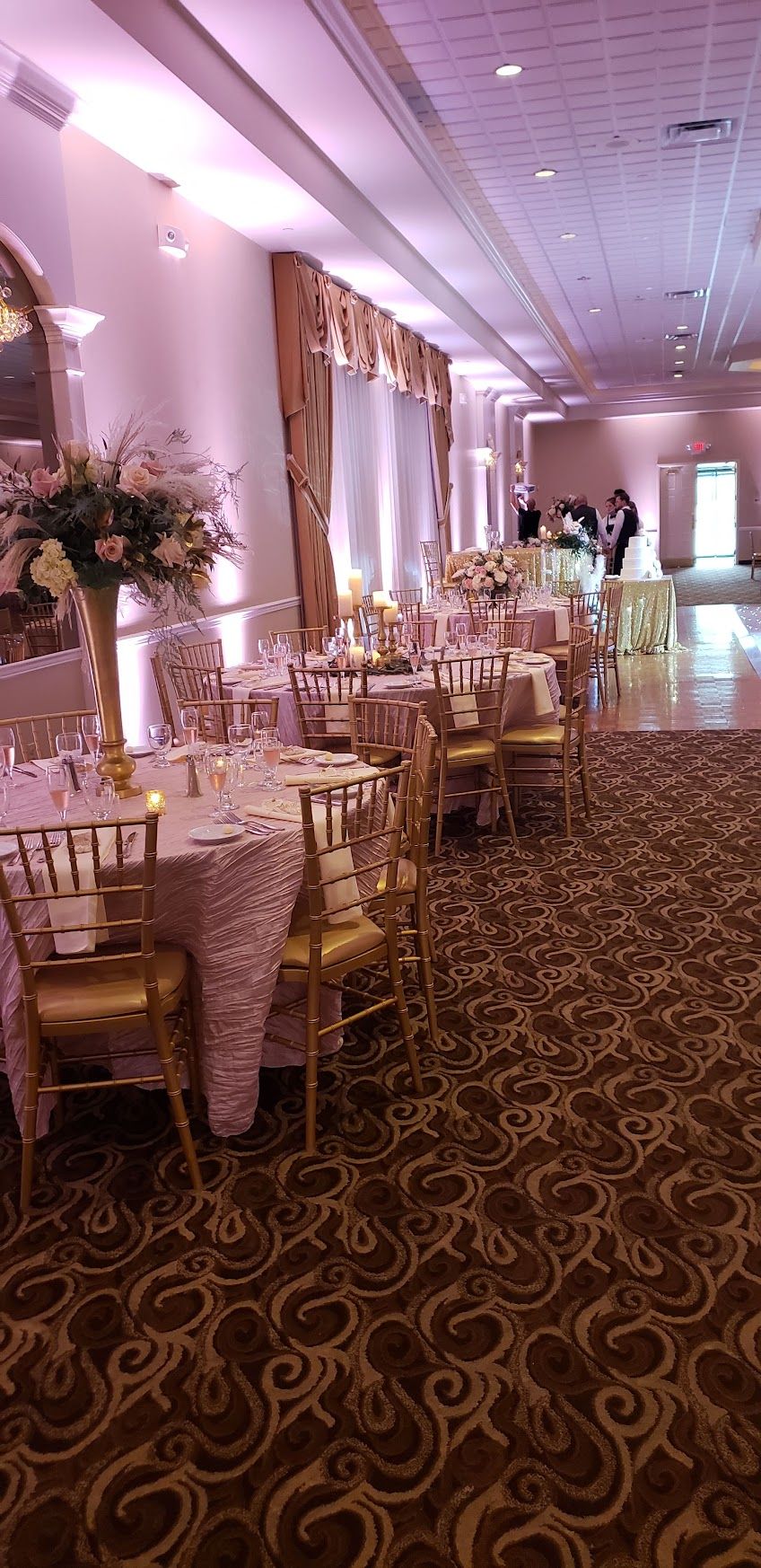 Elegant banquet hall with round tables, gold chairs, and pink-purple lighting along tall curtained windows
