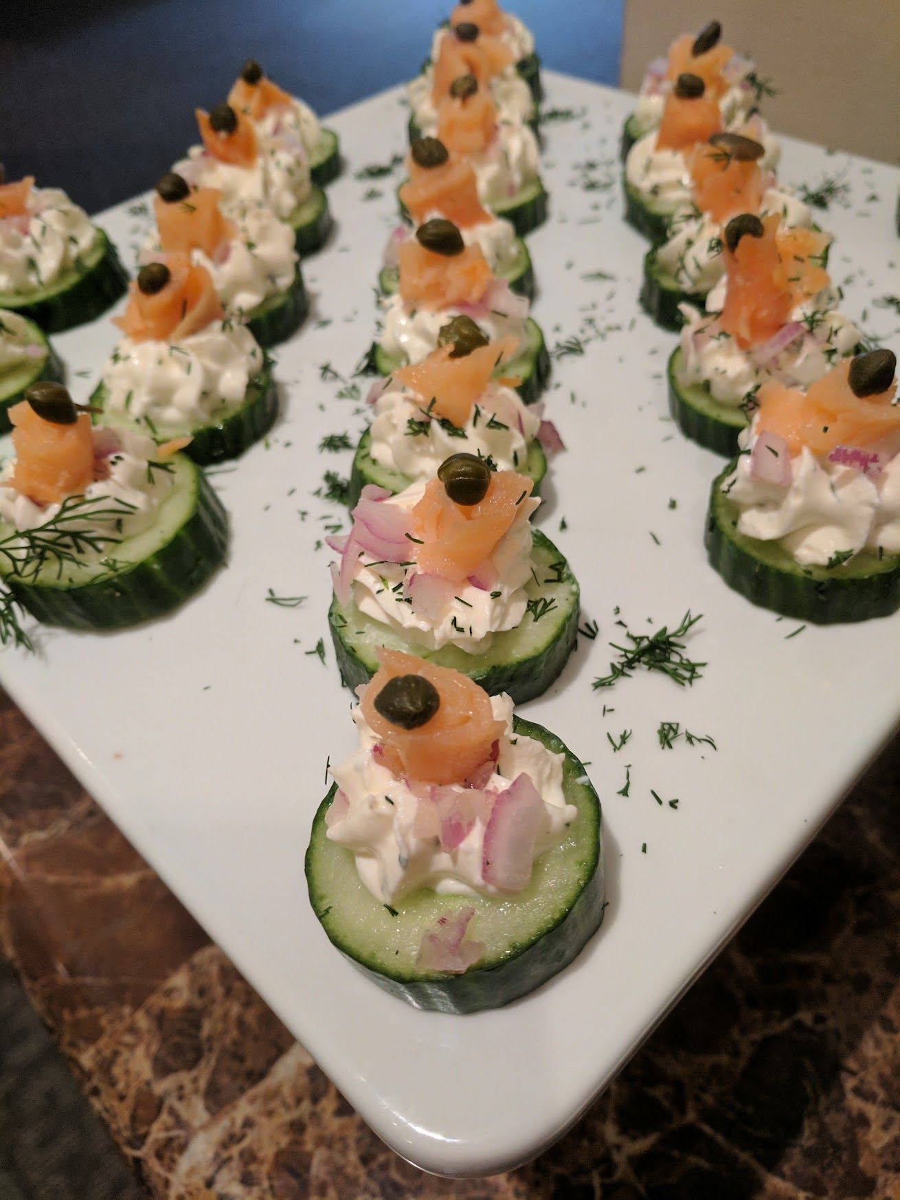 Platter of cucumber canapés topped with creamy salmon and herbs, arranged in neat rows on a white plate