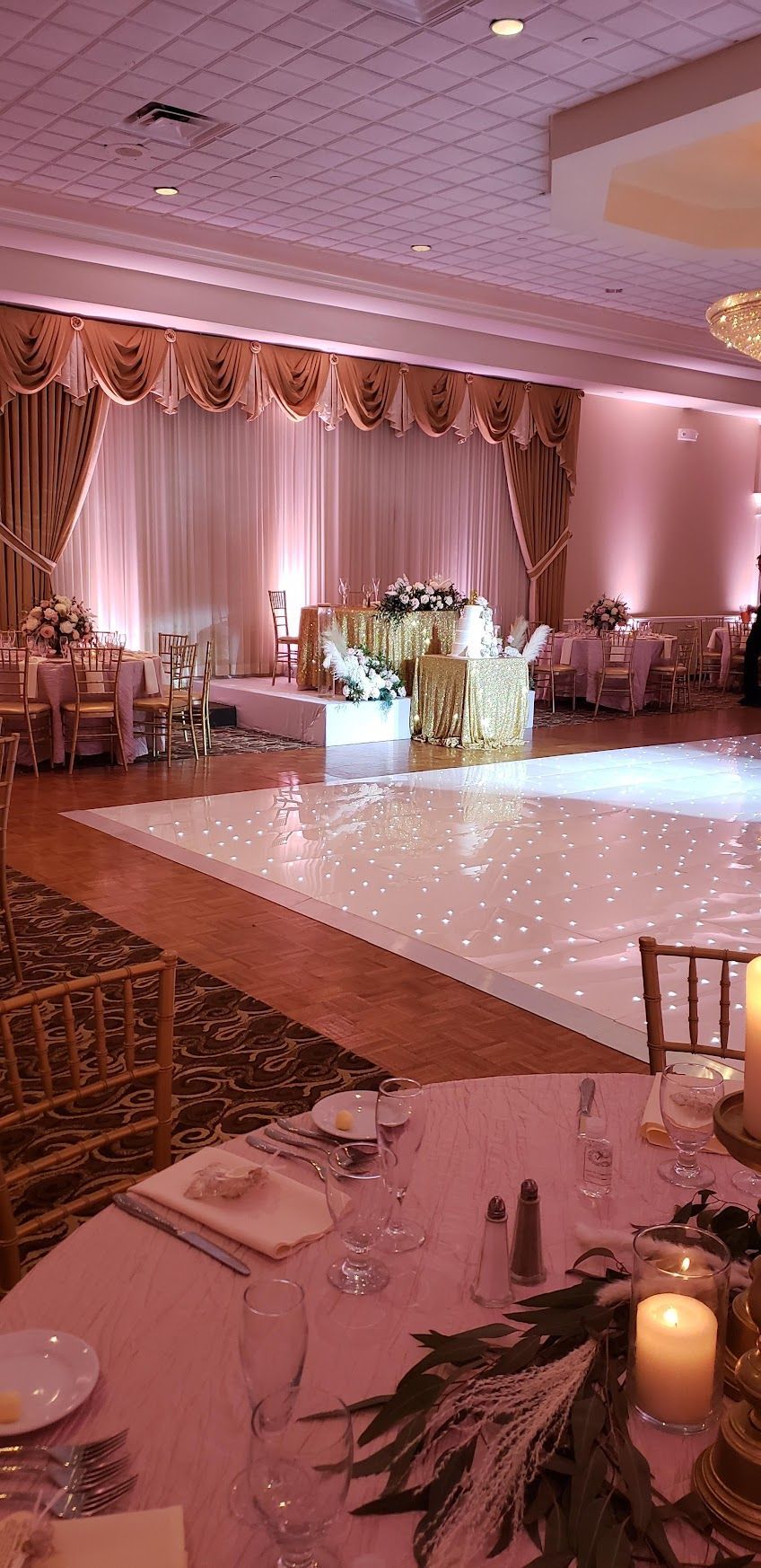 Elegant wedding reception hall with pink lighting, dance floor, round tables, and draped stage backdrop