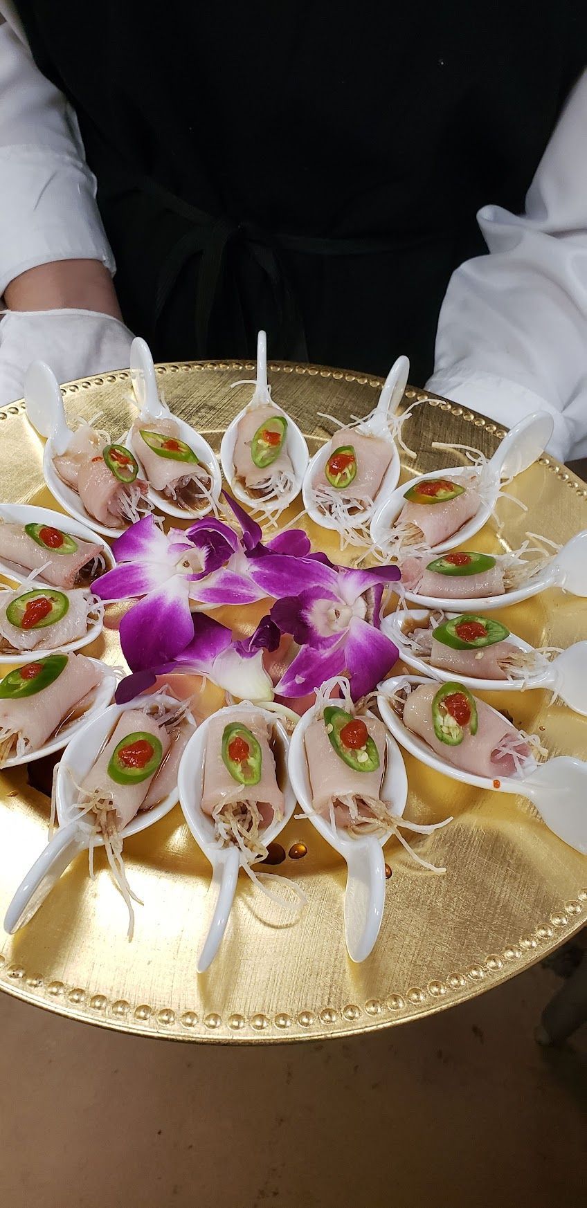 Gold tray of decorated white sweets topped with purple flowers and pearl-like garnish, held by two people