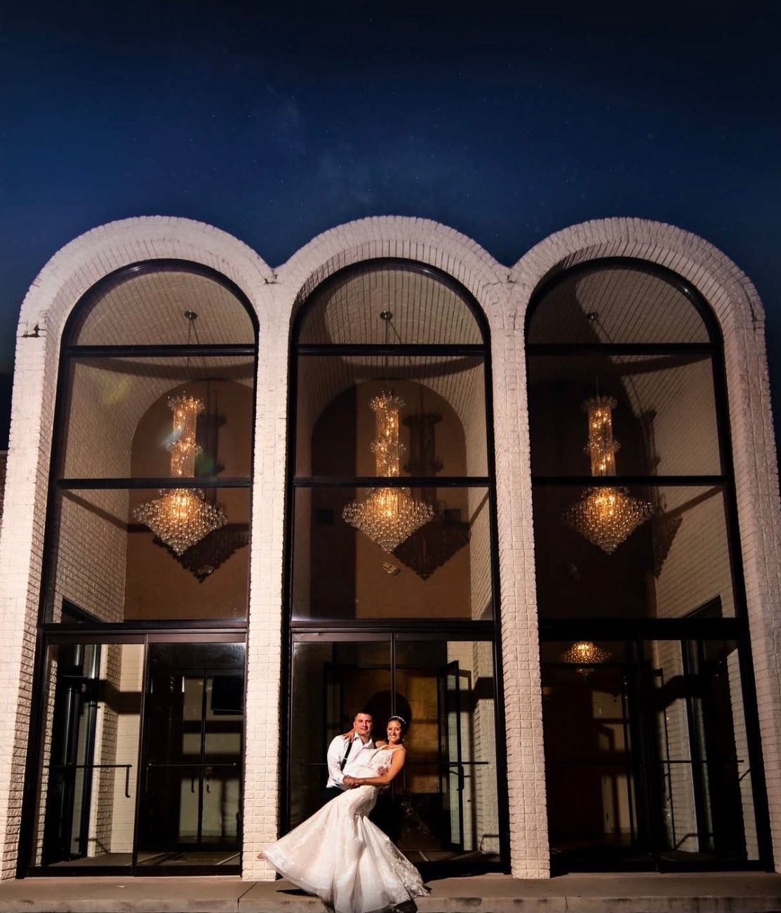 Bride and groom posing at the entrance of a building with arched windows at night