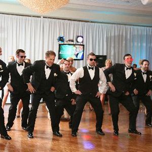 Several men in black vests and white shirts dance in formation at a formal indoor event.