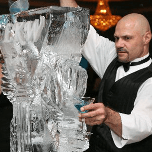 Bartender carving a clear ice sculpture while holding a blue drink in a formal setting