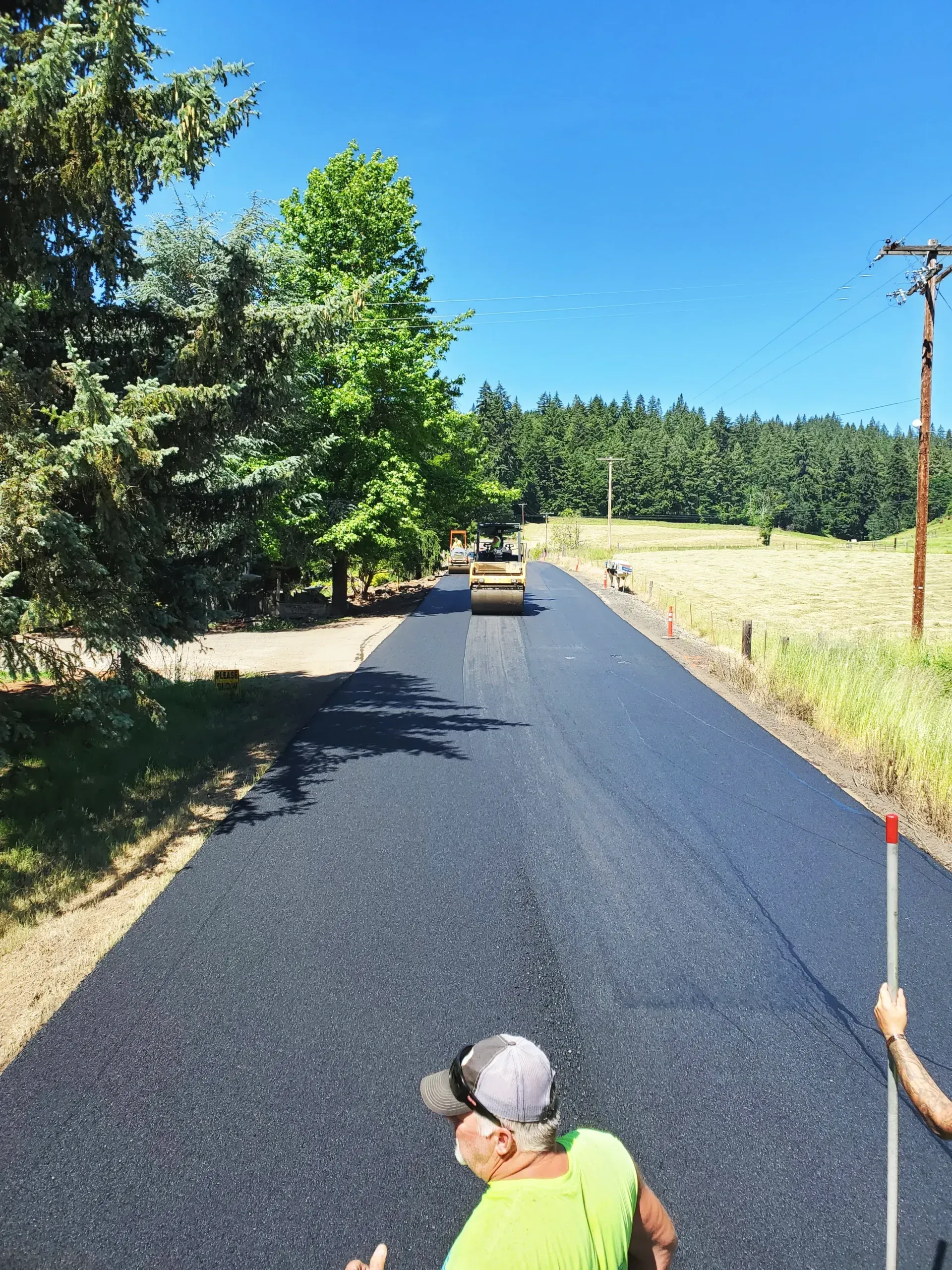 A worker in a high-visibility yellow shirt stands on a freshly paved asphalt road as a steamroller approaches in the sun.