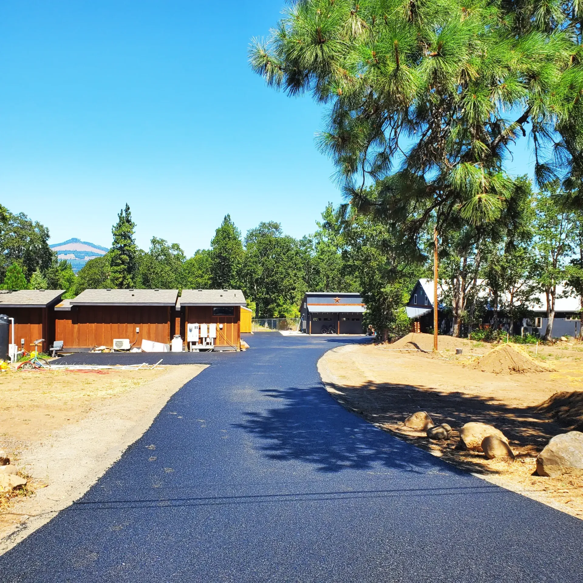 A freshly paved black asphalt driveway leads through a sunny, tree-lined property toward several wooden buildings.
