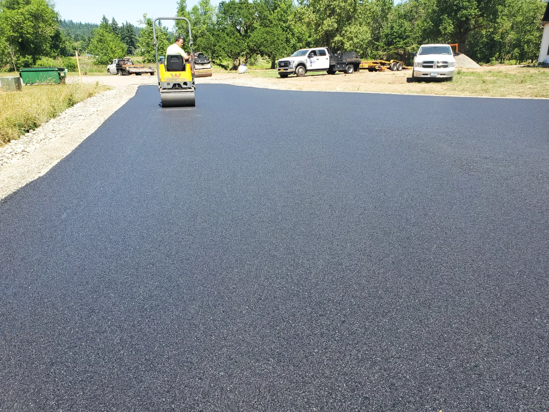 A small yellow roller compacts a freshly laid black asphalt driveway under a clear sky.