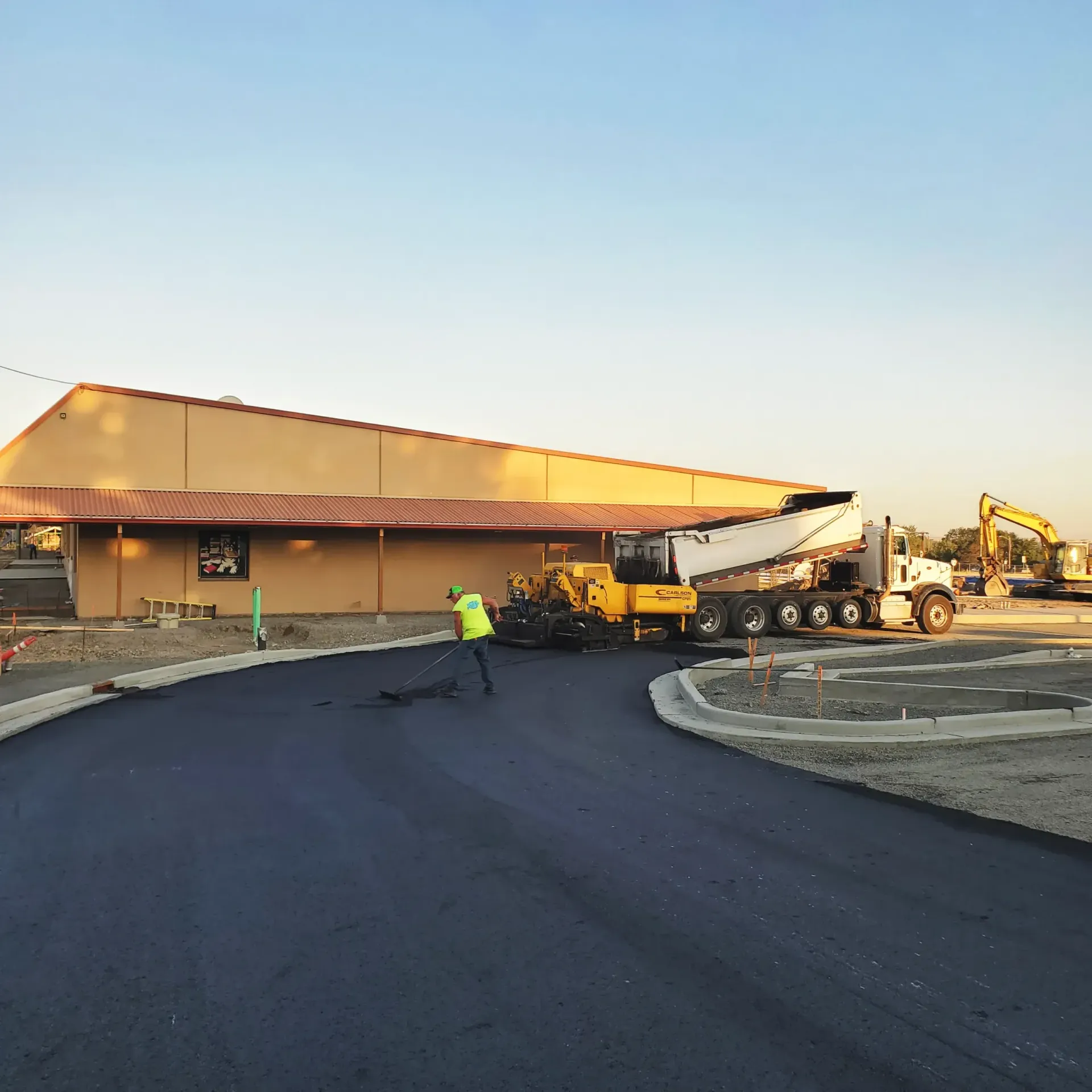 A worker in a yellow safety vest directs a paving machine laying asphalt in front of a tan commercial building.