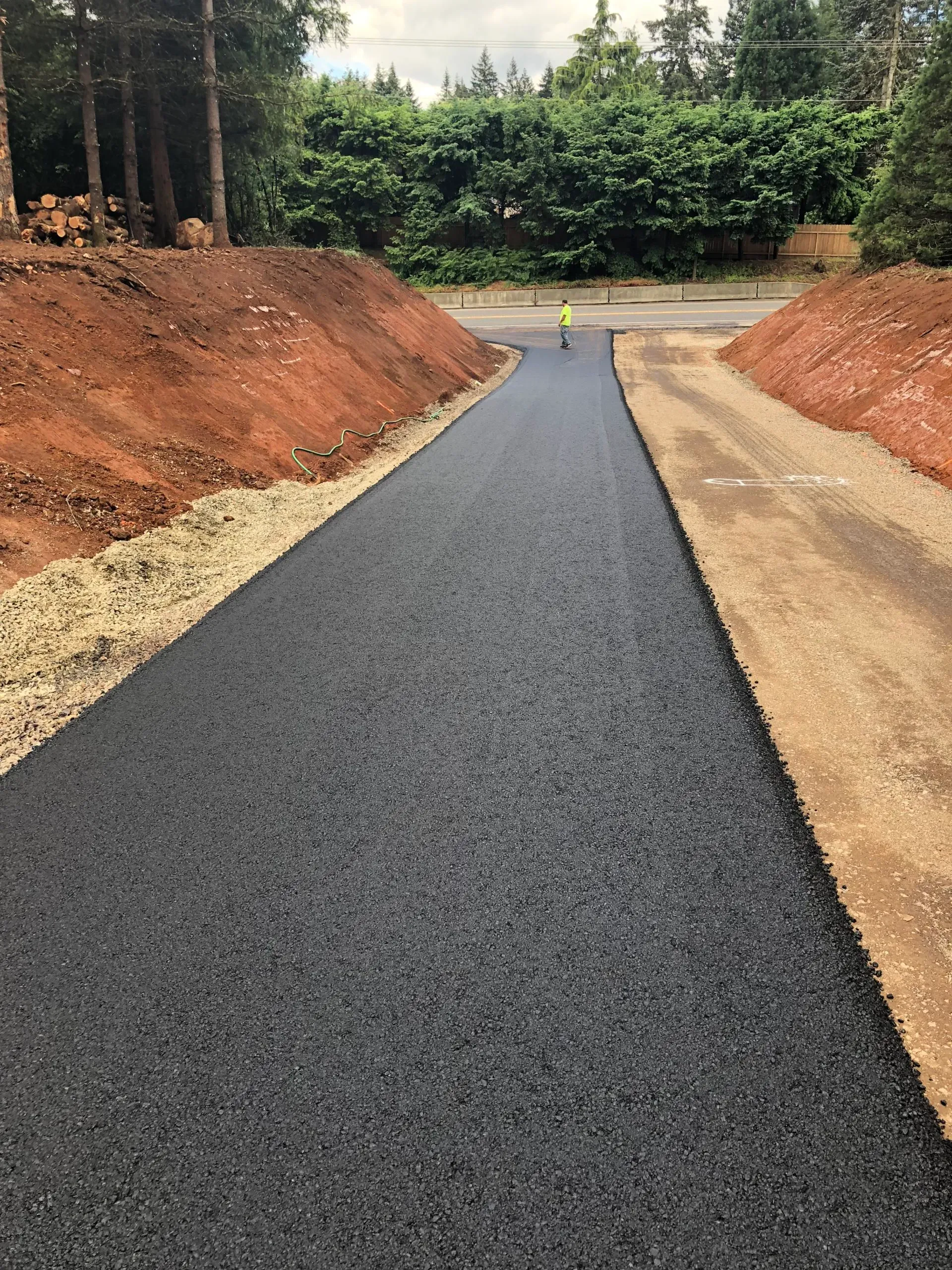 A freshly paved asphalt driveway leading toward trees, bordered by banks of reddish-brown soil and a gravel path.