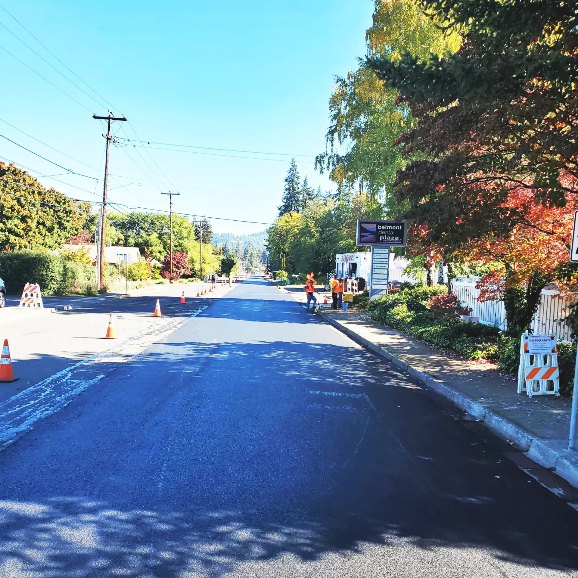 A freshly paved road under a blue sky, with construction workers on the side and traffic cones marking the area.