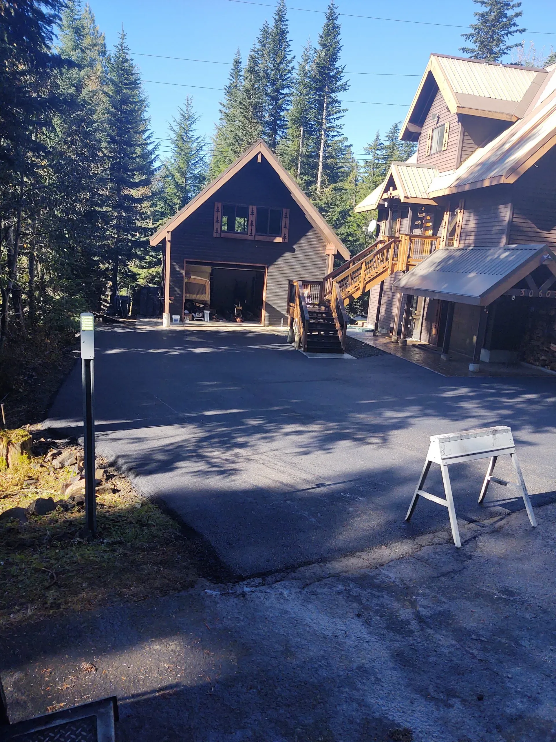 A dark wood garage and house exterior with a newly paved driveway and a white sawhorse in a wooded, sunny setting.