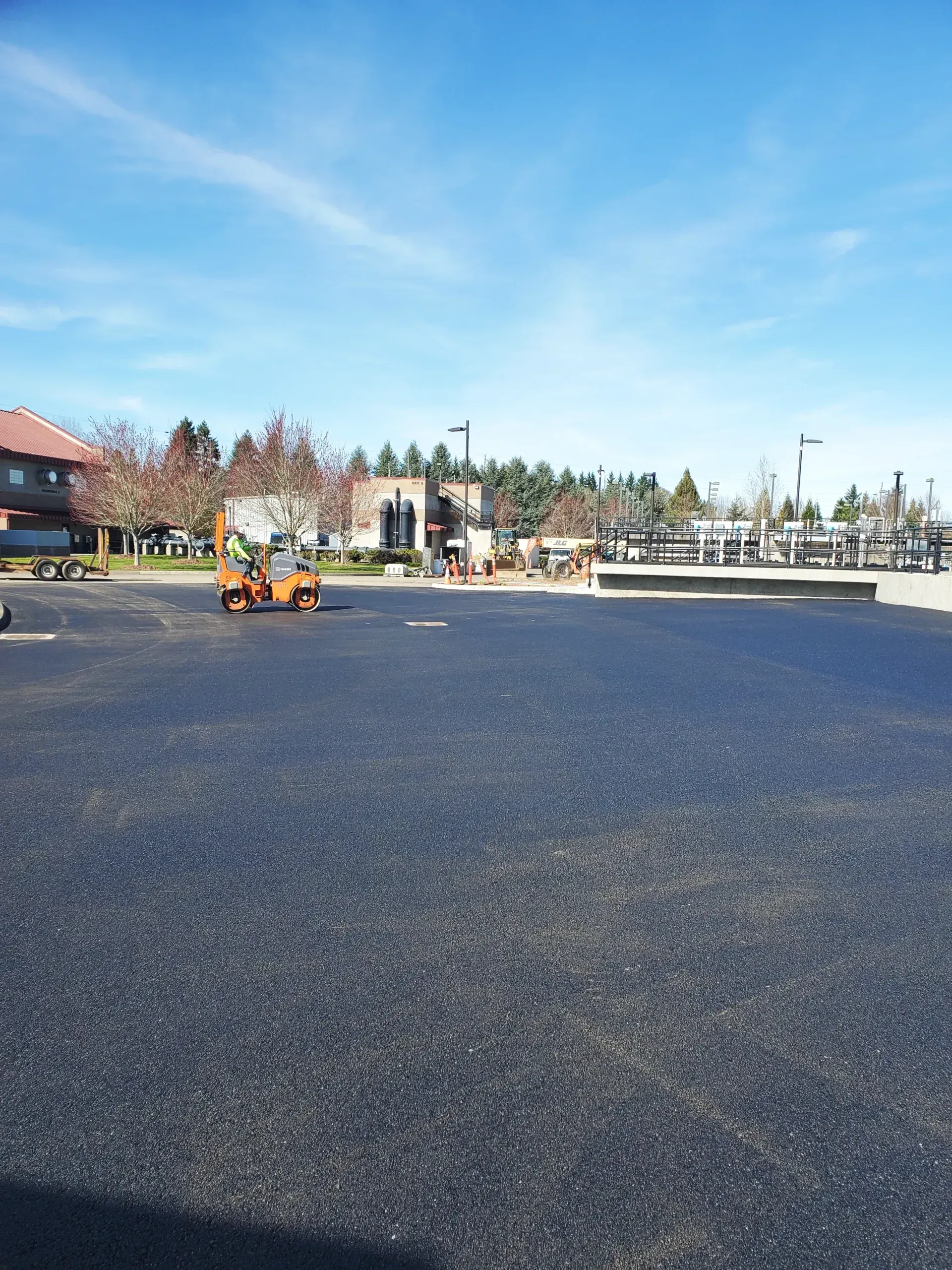 A small orange construction vehicle sits on a freshly paved, dark asphalt parking lot under a clear blue sky.