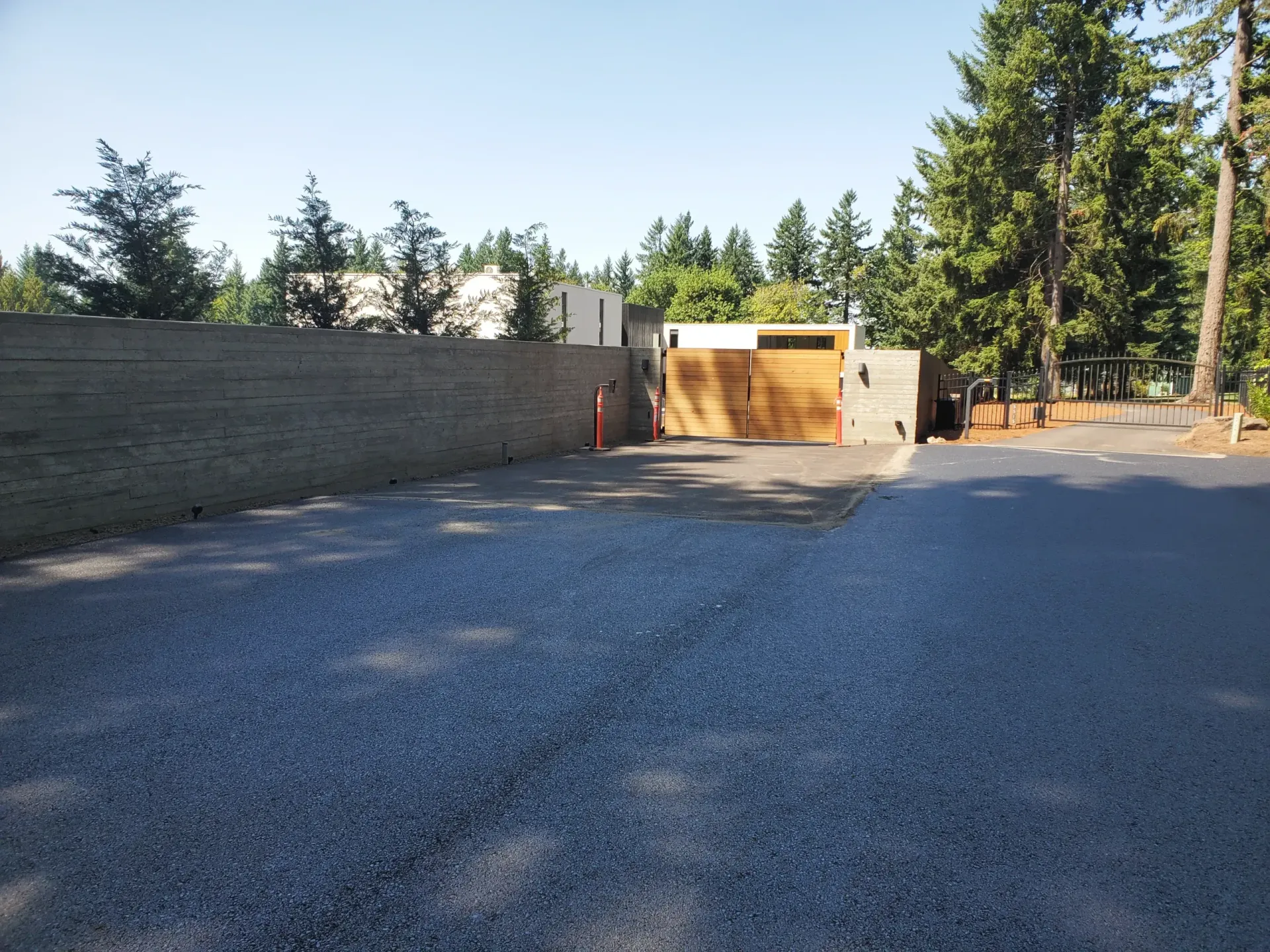 A paved driveway leads to a wooden gate and wall alongside trees under a clear blue sky.