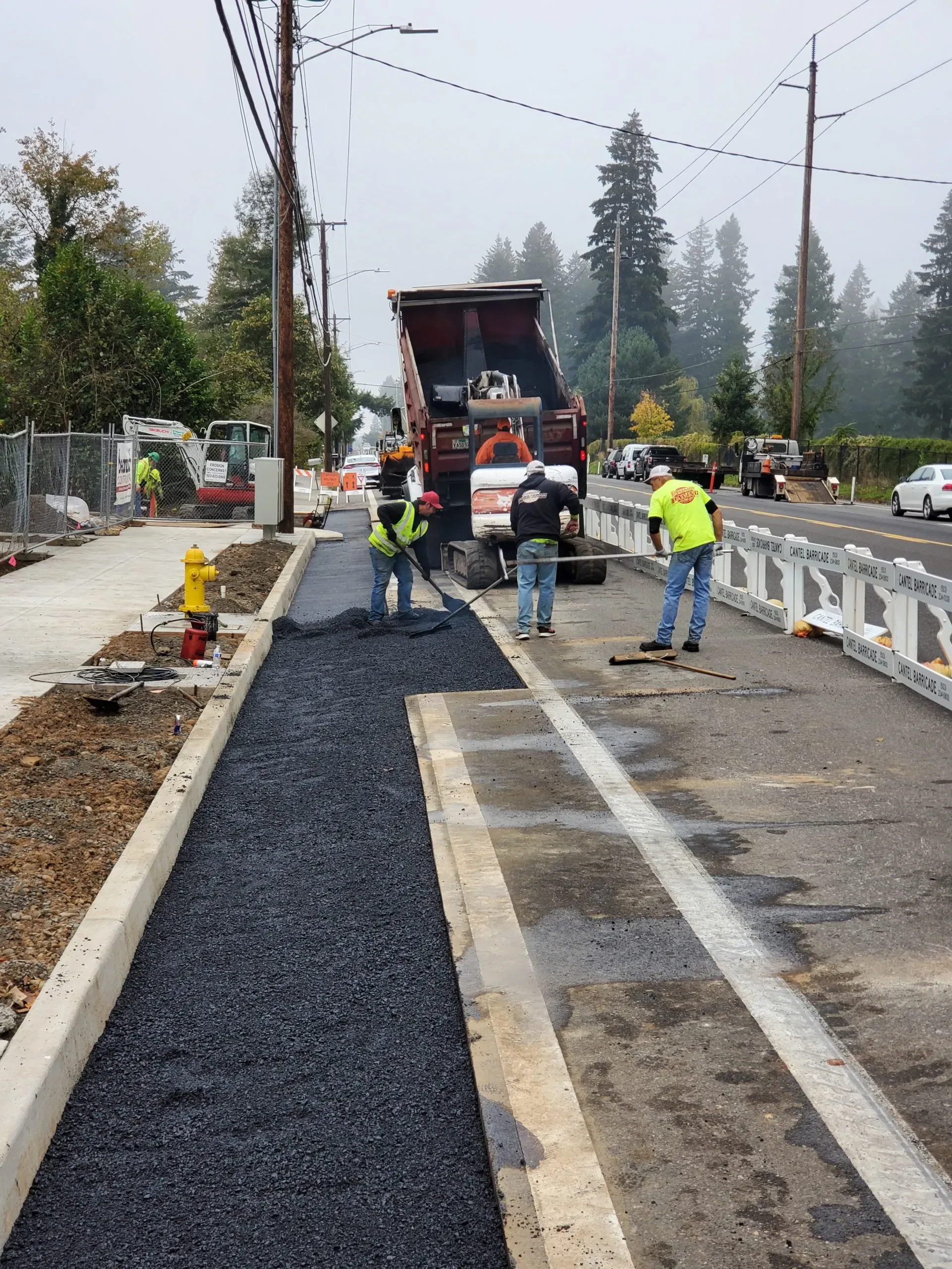 Three workers in safety gear install fresh asphalt on a sidewalk alongside a road with traffic barriers and a dump truck.