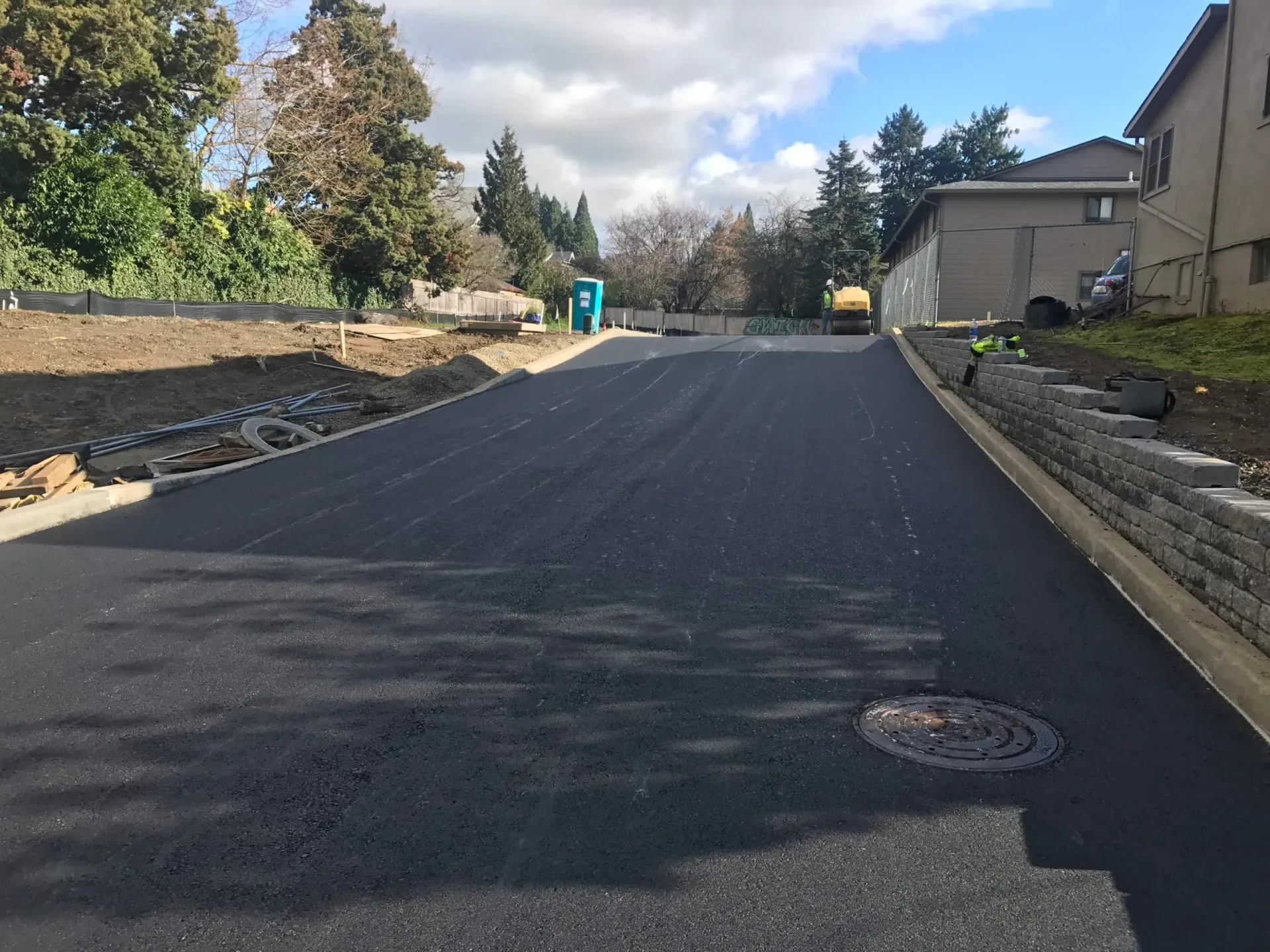 A freshly paved asphalt road leads toward a construction site with a blue portable toilet under a cloudy sky.
