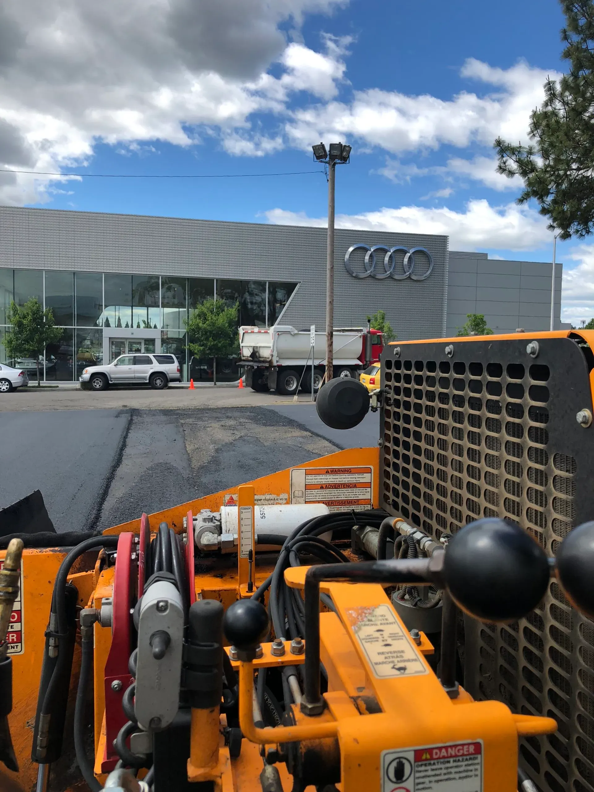 A view from the operator's seat of yellow heavy machinery facing an Audi dealership with a parked truck nearby.
