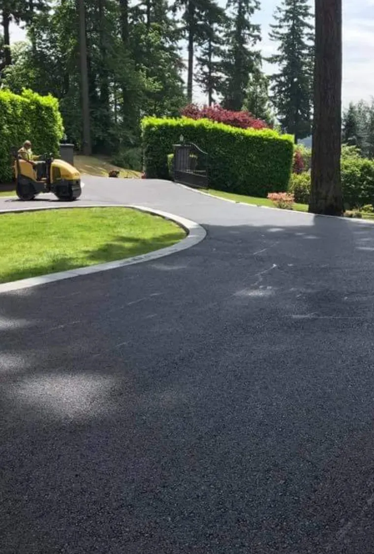 A yellow ride-on roller sits on a freshly paved dark asphalt driveway next to a grassy lawn and manicured hedges.