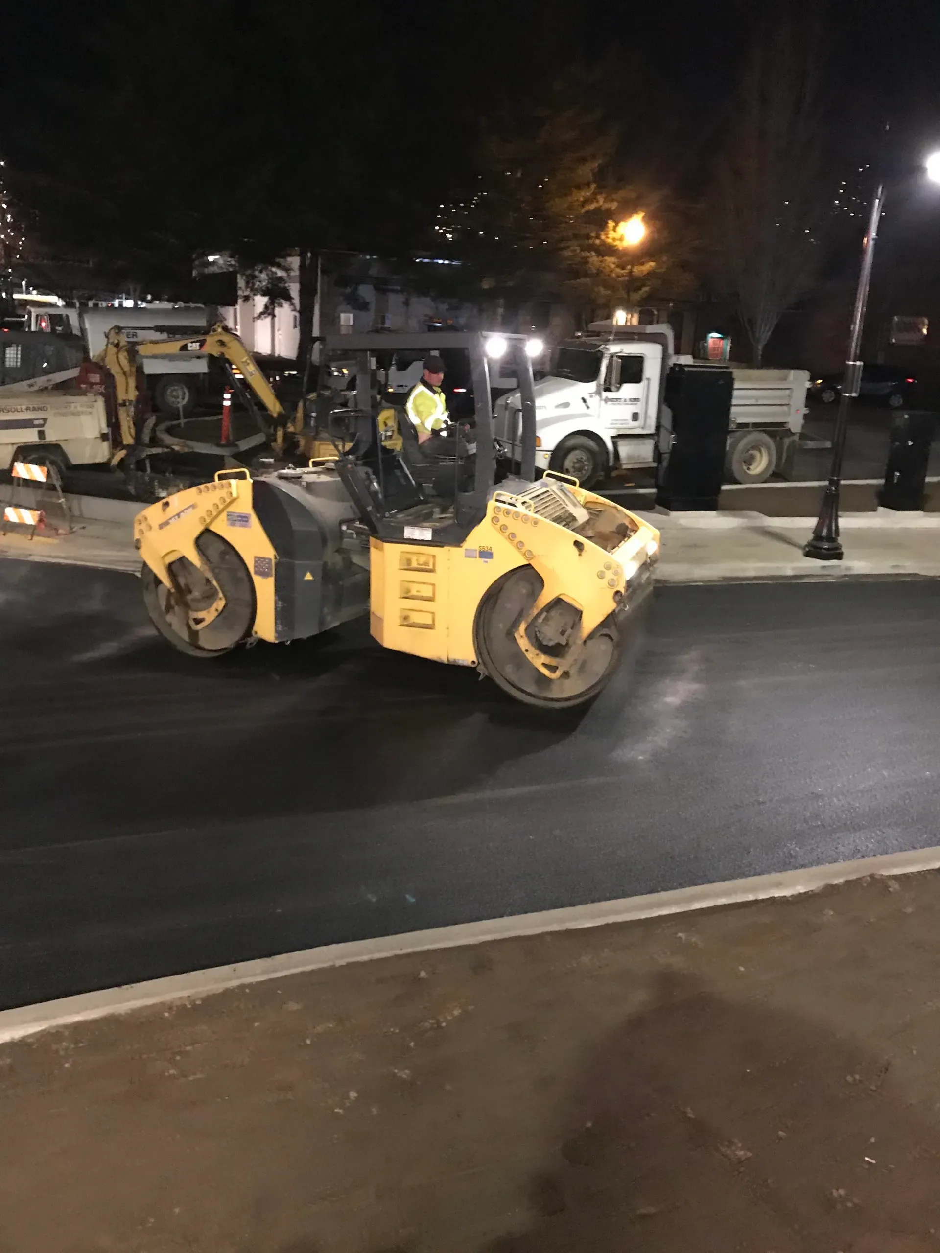 A yellow industrial road roller works on fresh, dark asphalt at night, with trucks parked in the background.