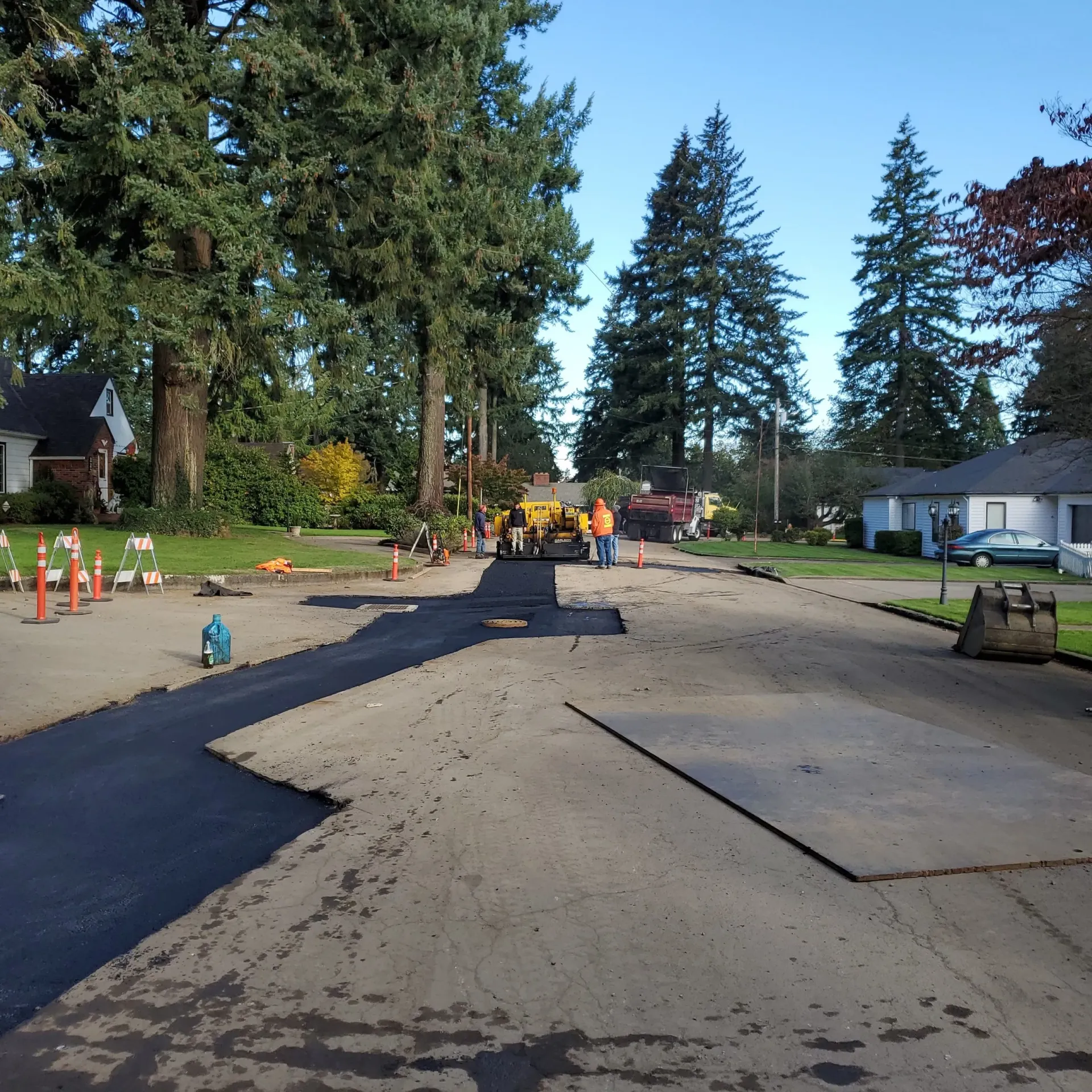 A road repair crew working on a suburban street, laying a new patch of black asphalt amid older, worn pavement.