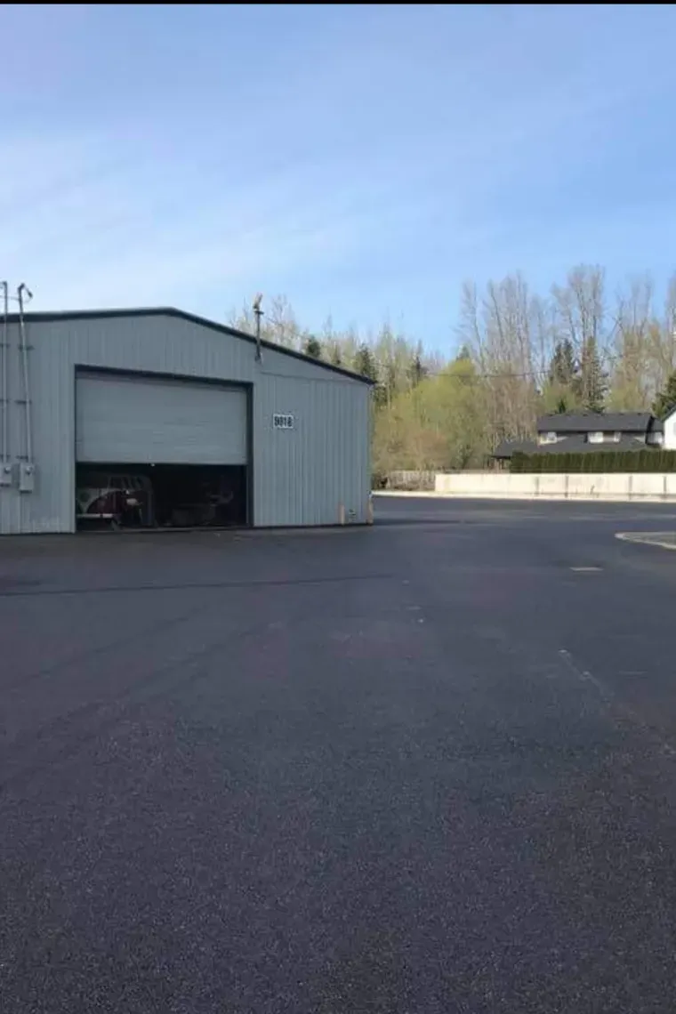 A light grey metal warehouse building with an open bay door stands on a paved lot under a clear blue sky.
