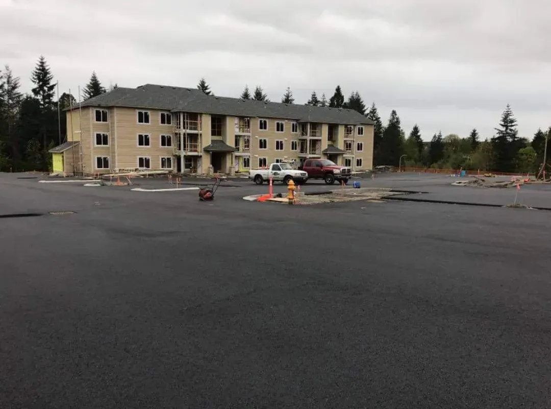 A three-story apartment building behind a large, recently paved black asphalt parking lot with construction markers.