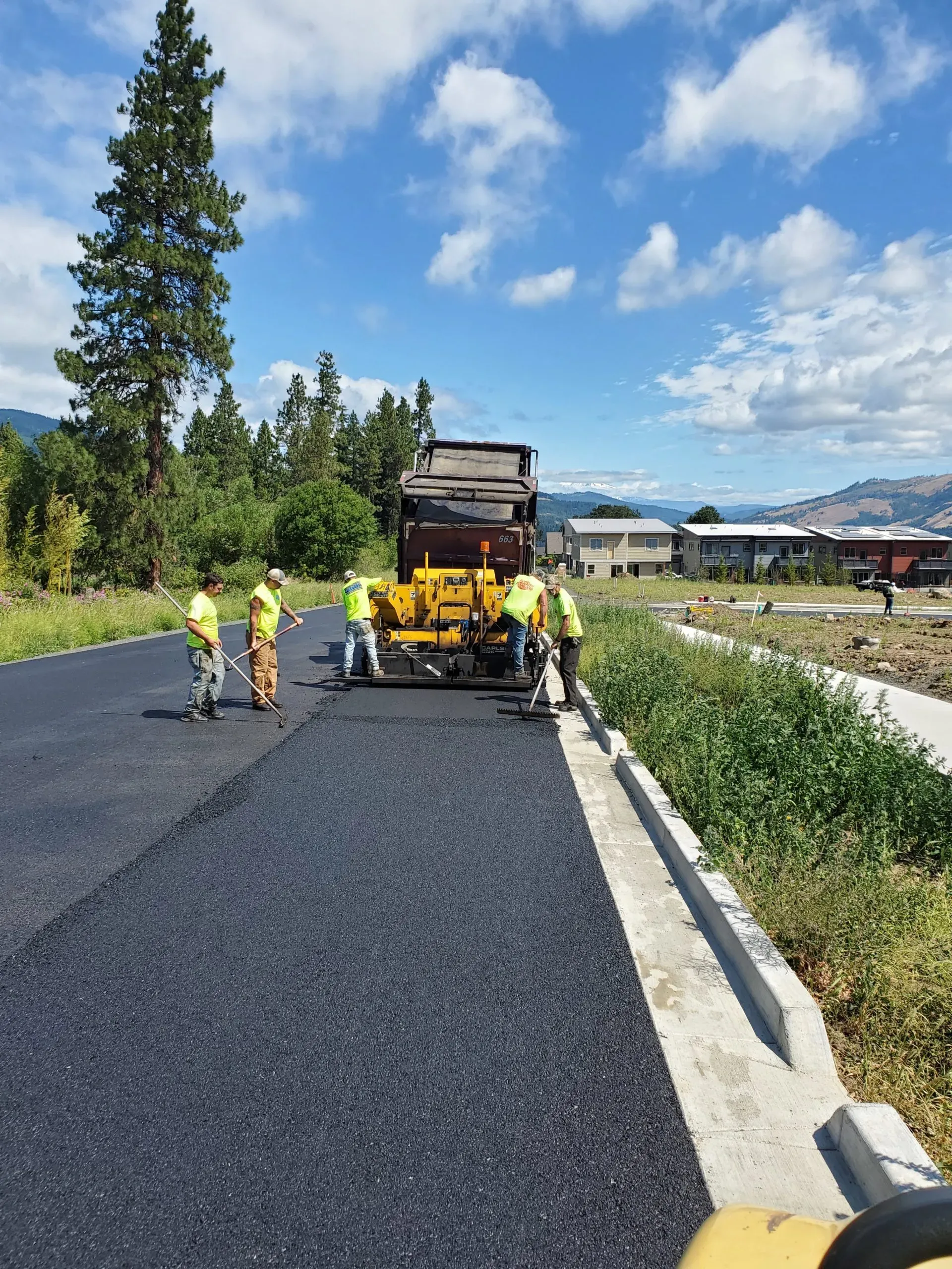 A road paving crew in neon safety vests operates machinery to lay fresh asphalt on a sunny, suburban street.