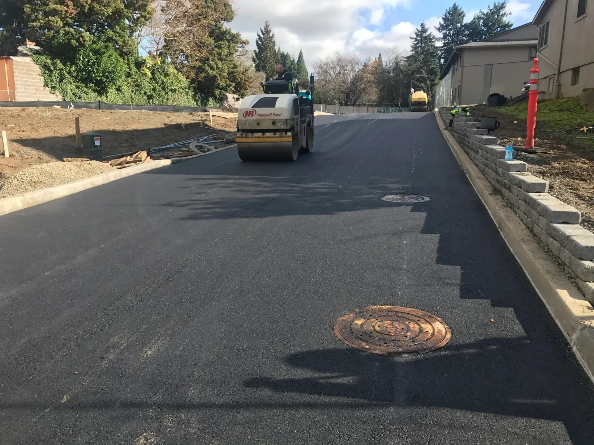 A construction vehicle rolls over a freshly paved asphalt road with a circular utility cover in the foreground.