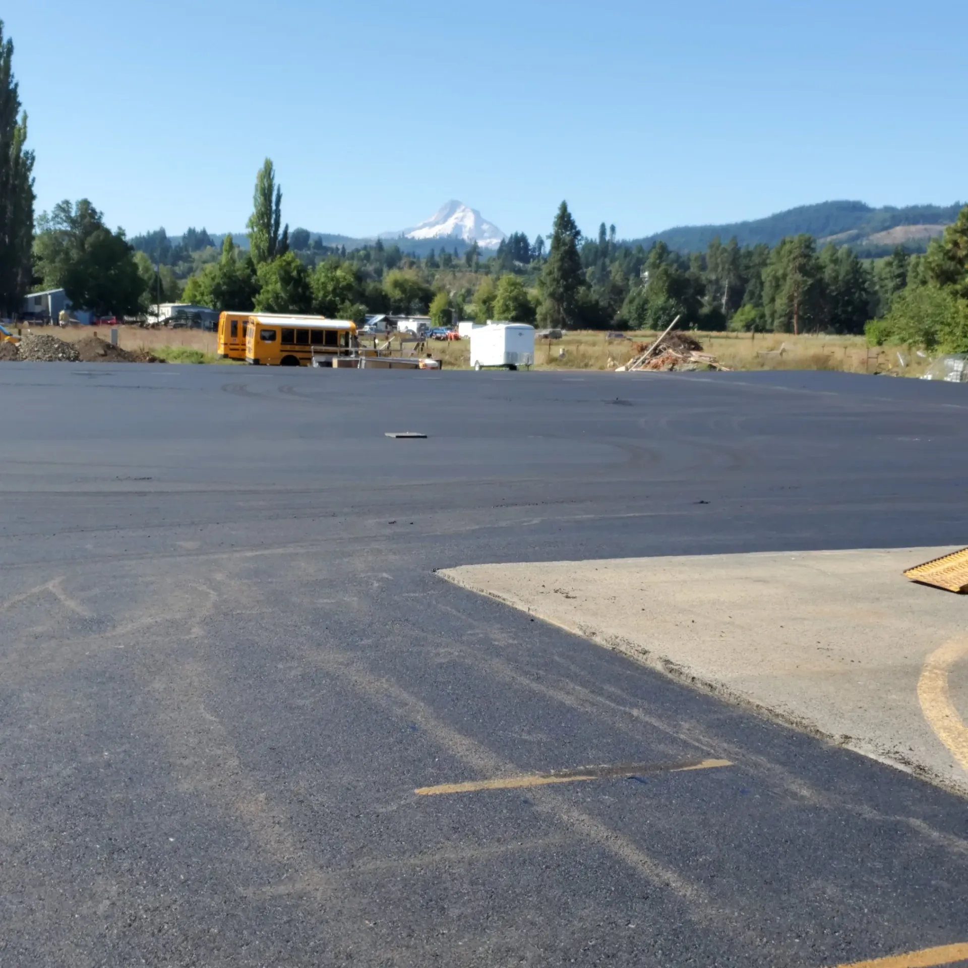 A large, freshly paved asphalt parking lot sits before a field with a yellow school bus, trees, and a snow-capped mountain.