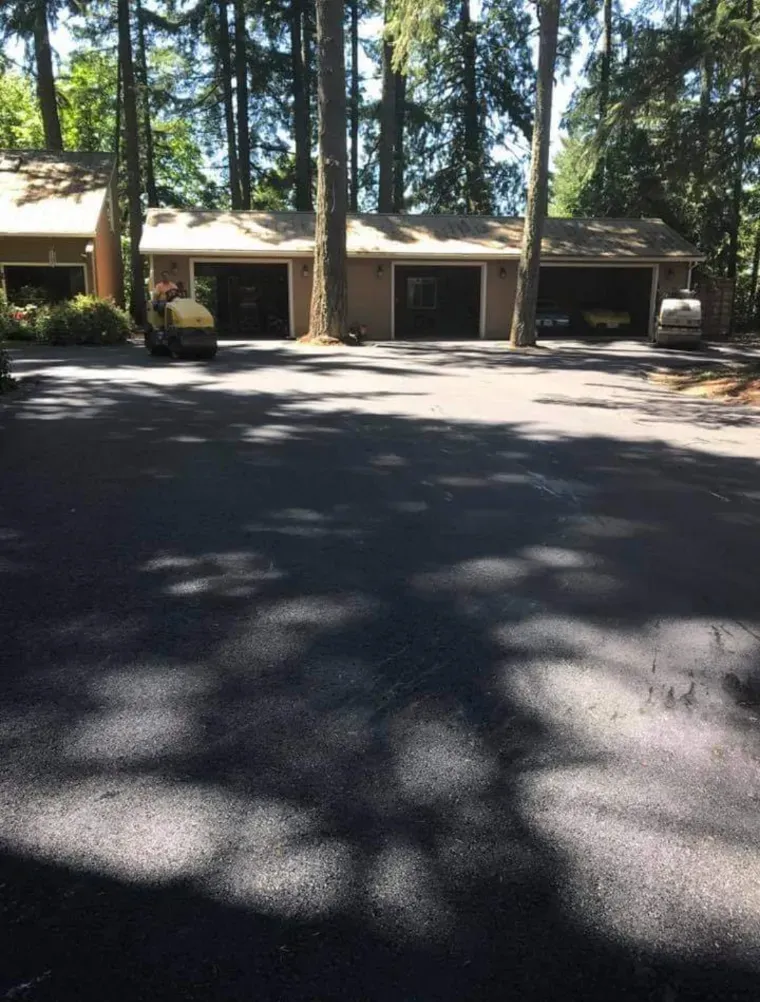 A freshly paved asphalt driveway leading to a three-car garage in a wooded, residential setting.