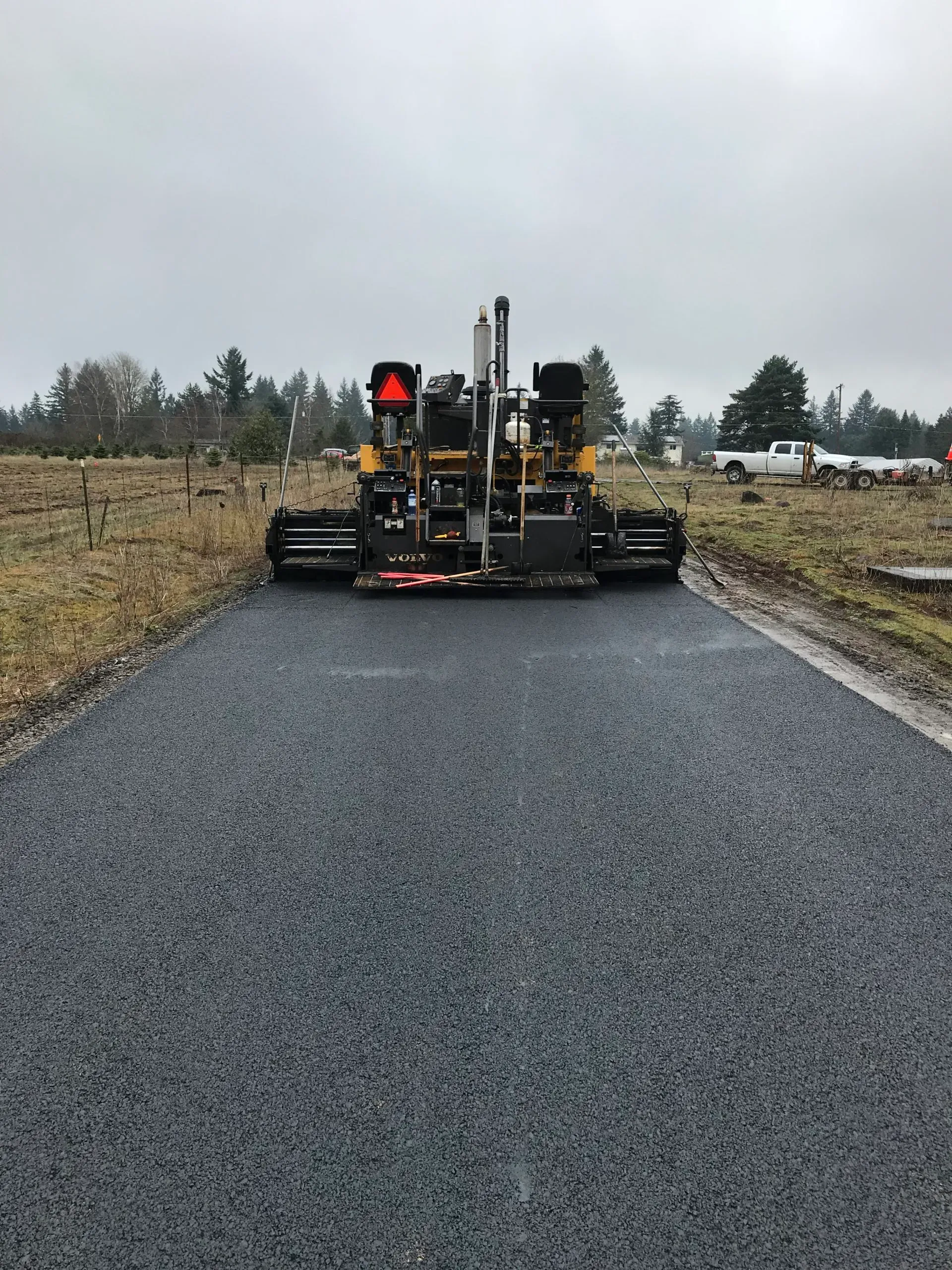 An asphalt paver laying down fresh black pavement on a dirt road under an overcast sky.