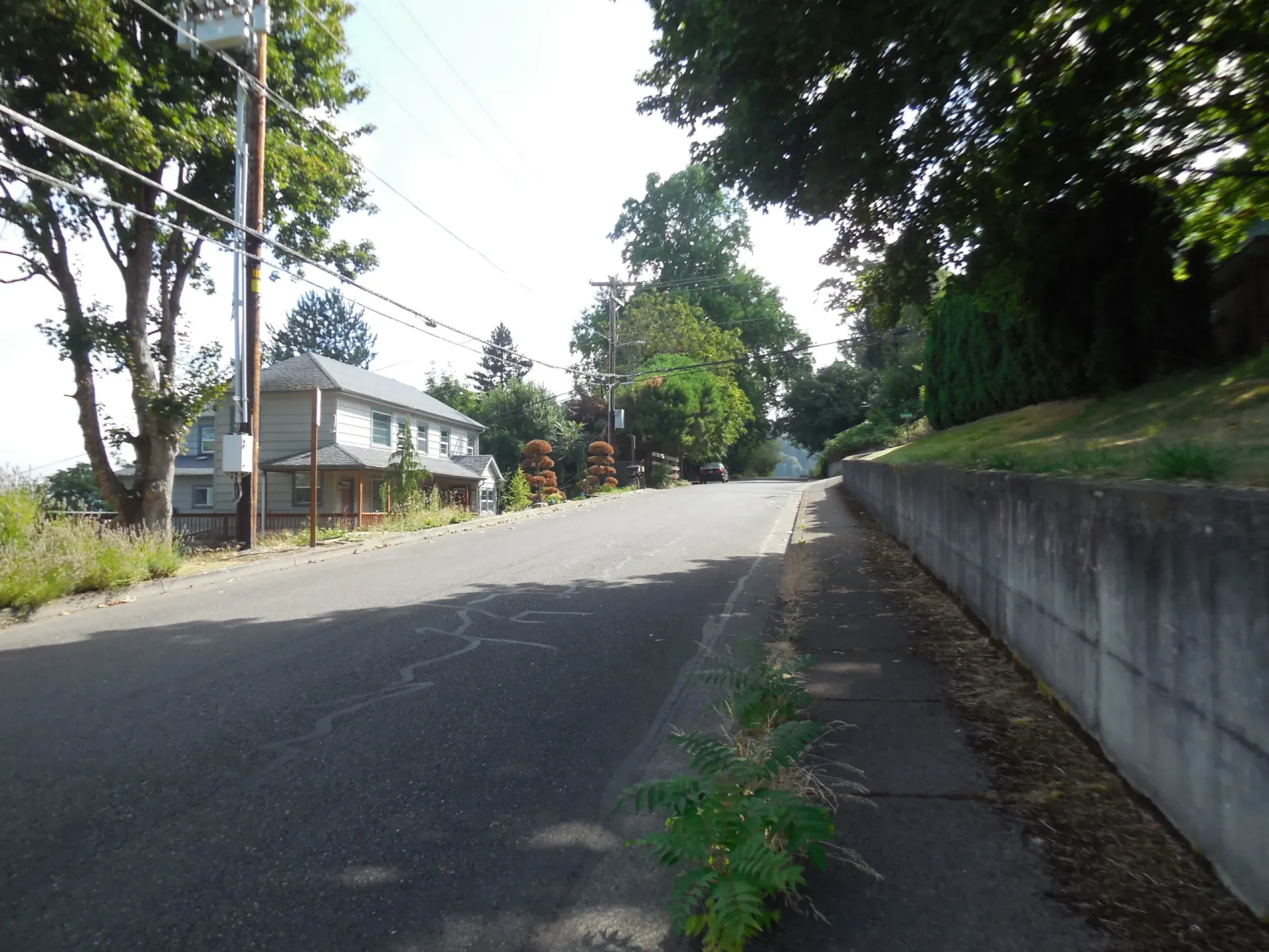 A paved road leads upward past a house on the left and a concrete retaining wall bordering a grassy bank on the right.