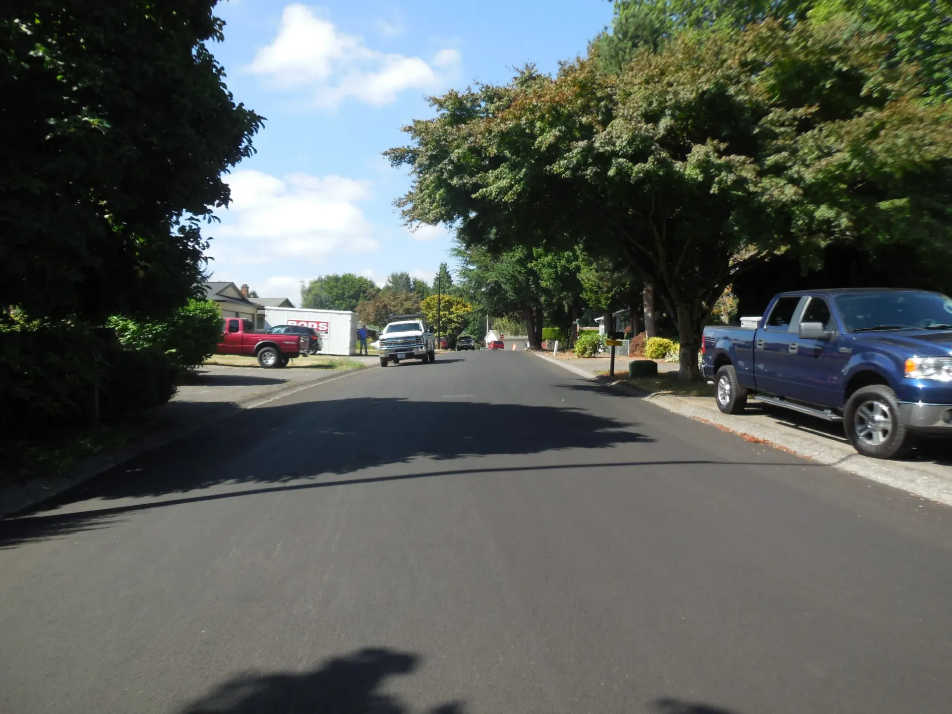 A paved residential street lined with trees, with several pickup trucks parked along the sides and in the distance.