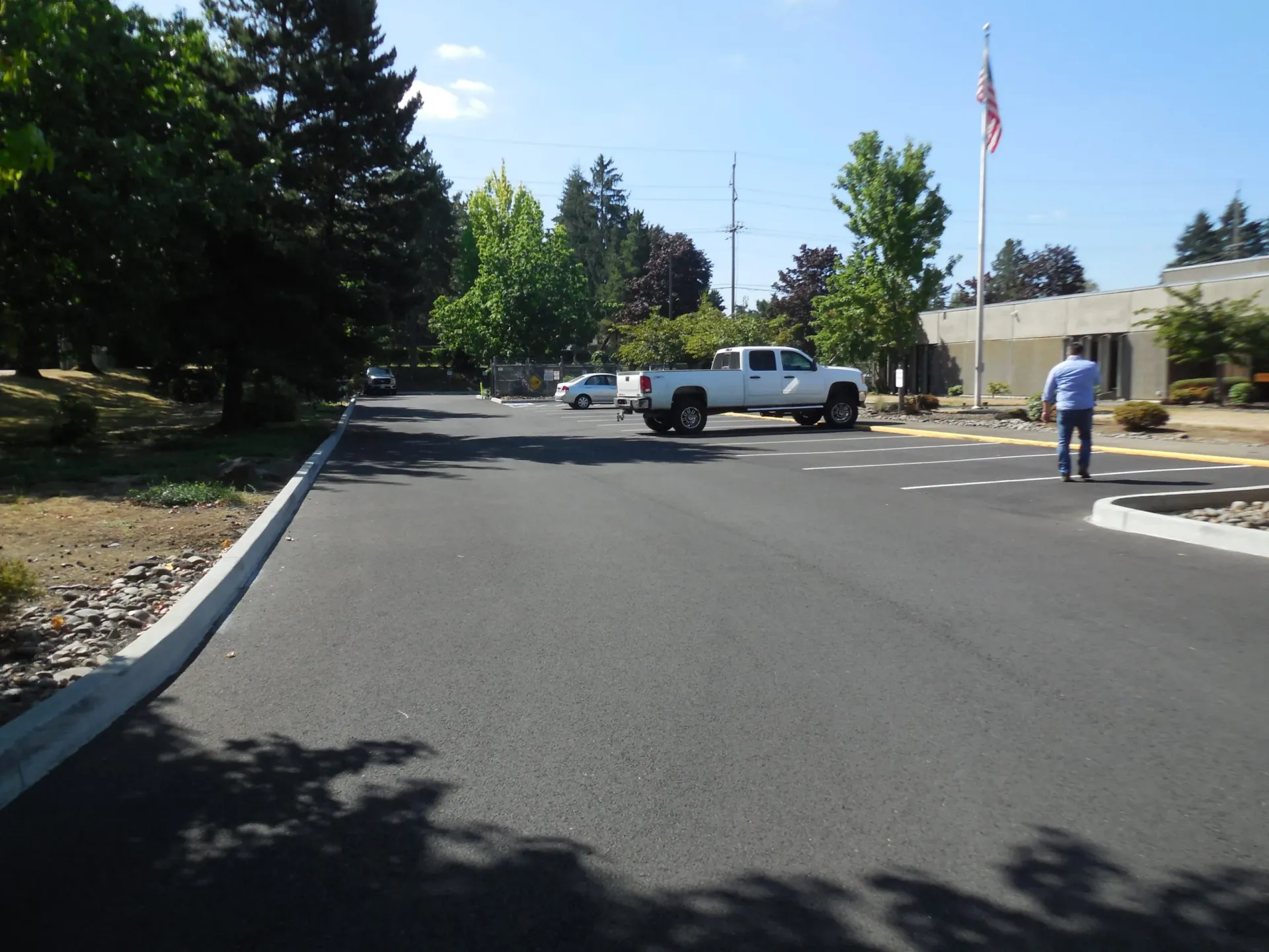 An asphalt parking lot on a sunny day with a white pickup truck, a person walking, and a flag pole.
