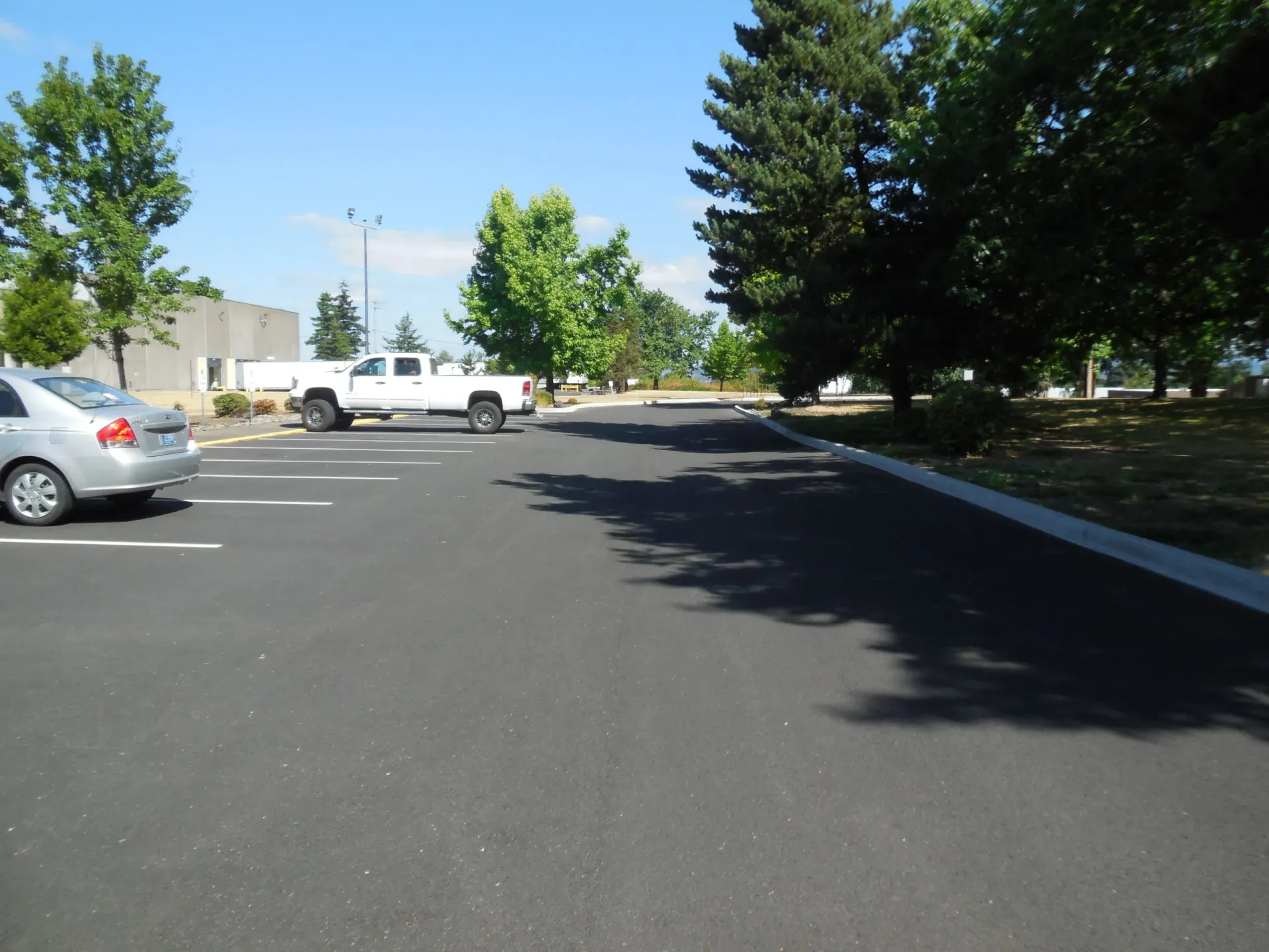 A paved parking lot under a clear blue sky, featuring a white pickup truck, a silver sedan, and surrounding green trees.