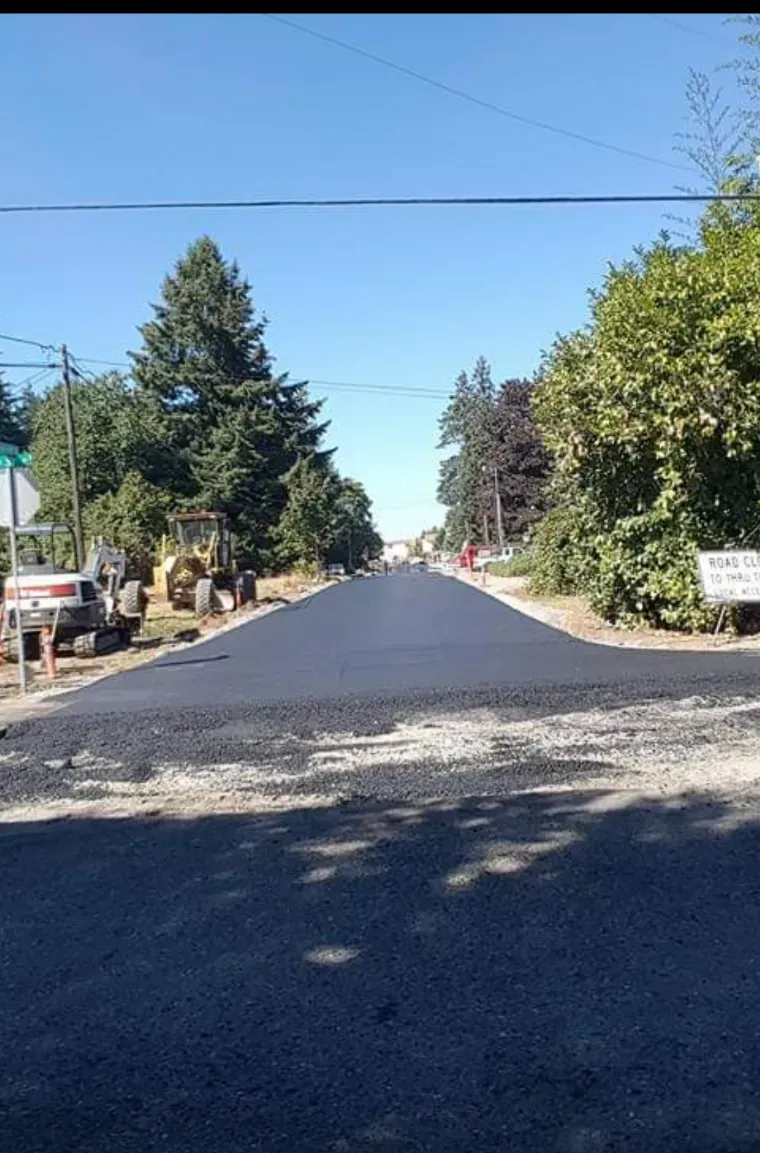 A newly paved black asphalt road stretches forward, flanked by trees and construction equipment on a sunny day.