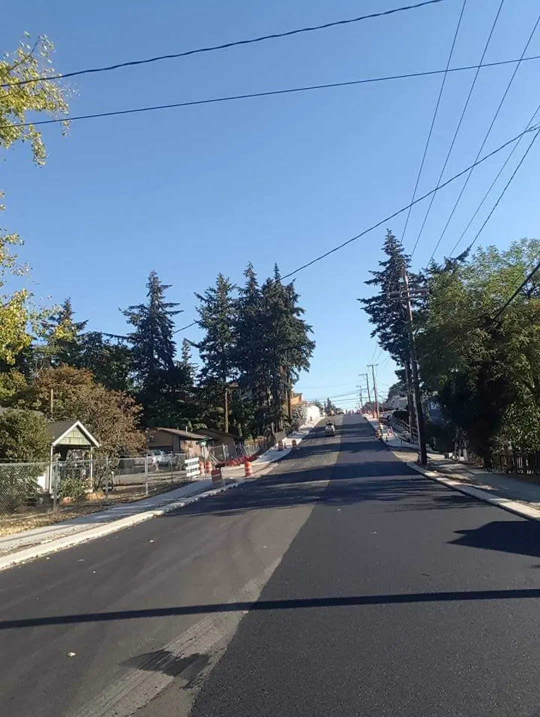 A freshly paved road stretches uphill under a clear blue sky, flanked by trees, sidewalks, and utility poles.