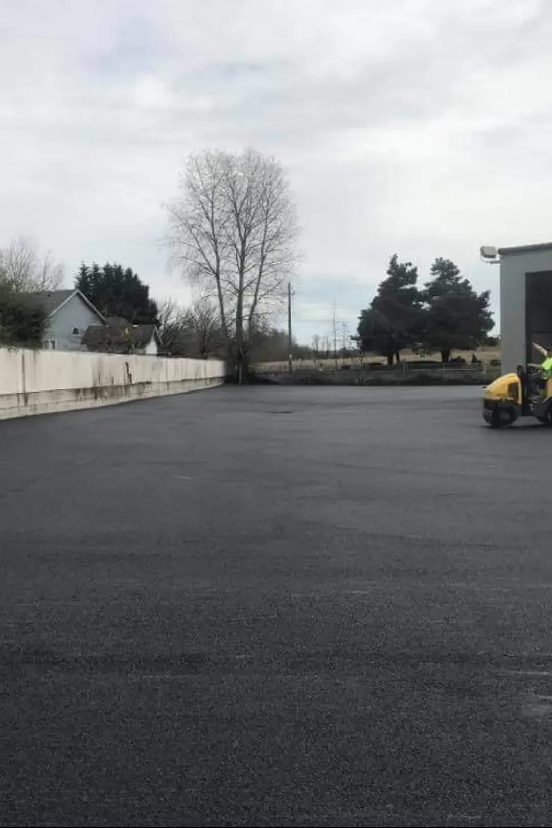 A construction worker operates a yellow asphalt roller on a newly paved parking lot next to a white concrete wall.