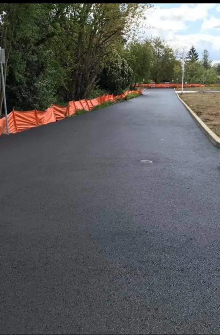 A newly paved asphalt path curves through a grassy area, lined with orange safety fencing and a concrete curb.