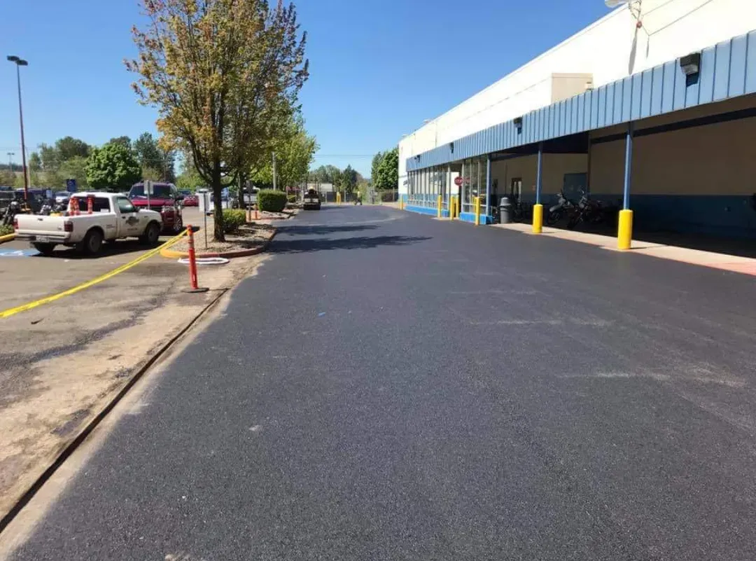 A freshly paved dark asphalt driveway leads alongside a large building with a blue covered walkway on a sunny day.