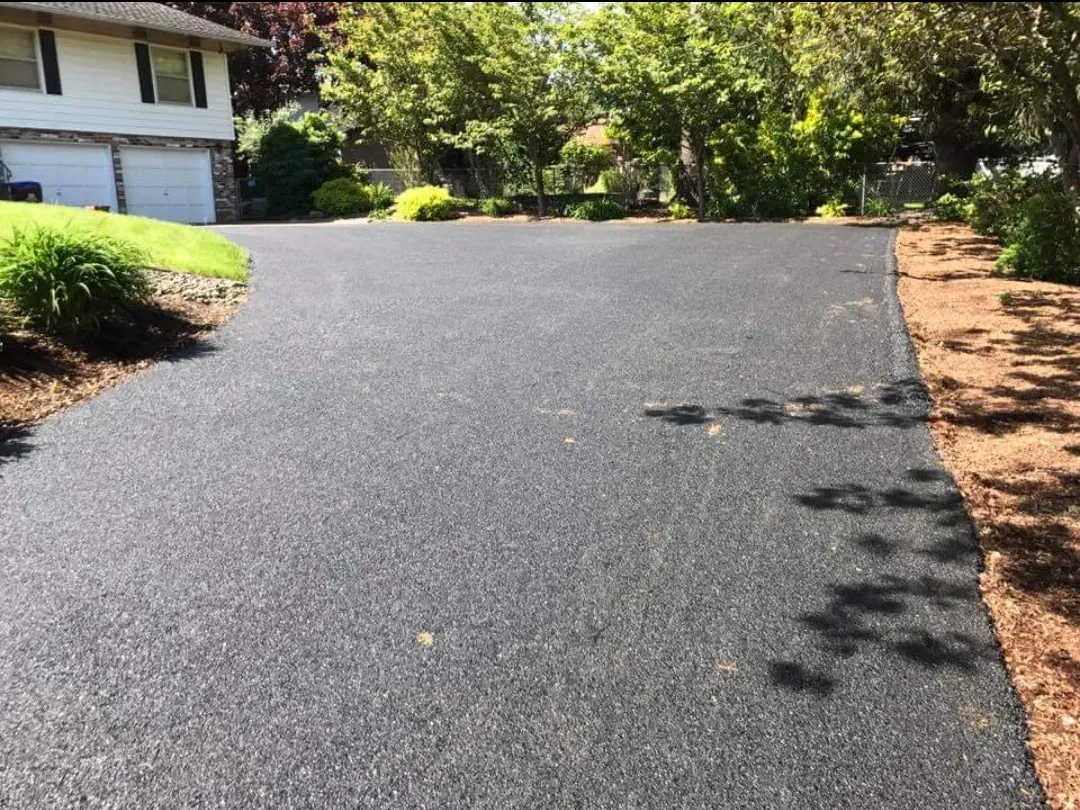 A freshly paved dark asphalt driveway leads to a house with white walls, flanked by green trees and landscaped beds.