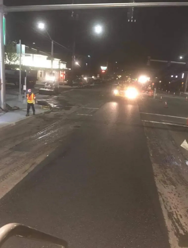 A worker in a high-visibility vest directs traffic at night near a road construction site with bright vehicle headlights.
