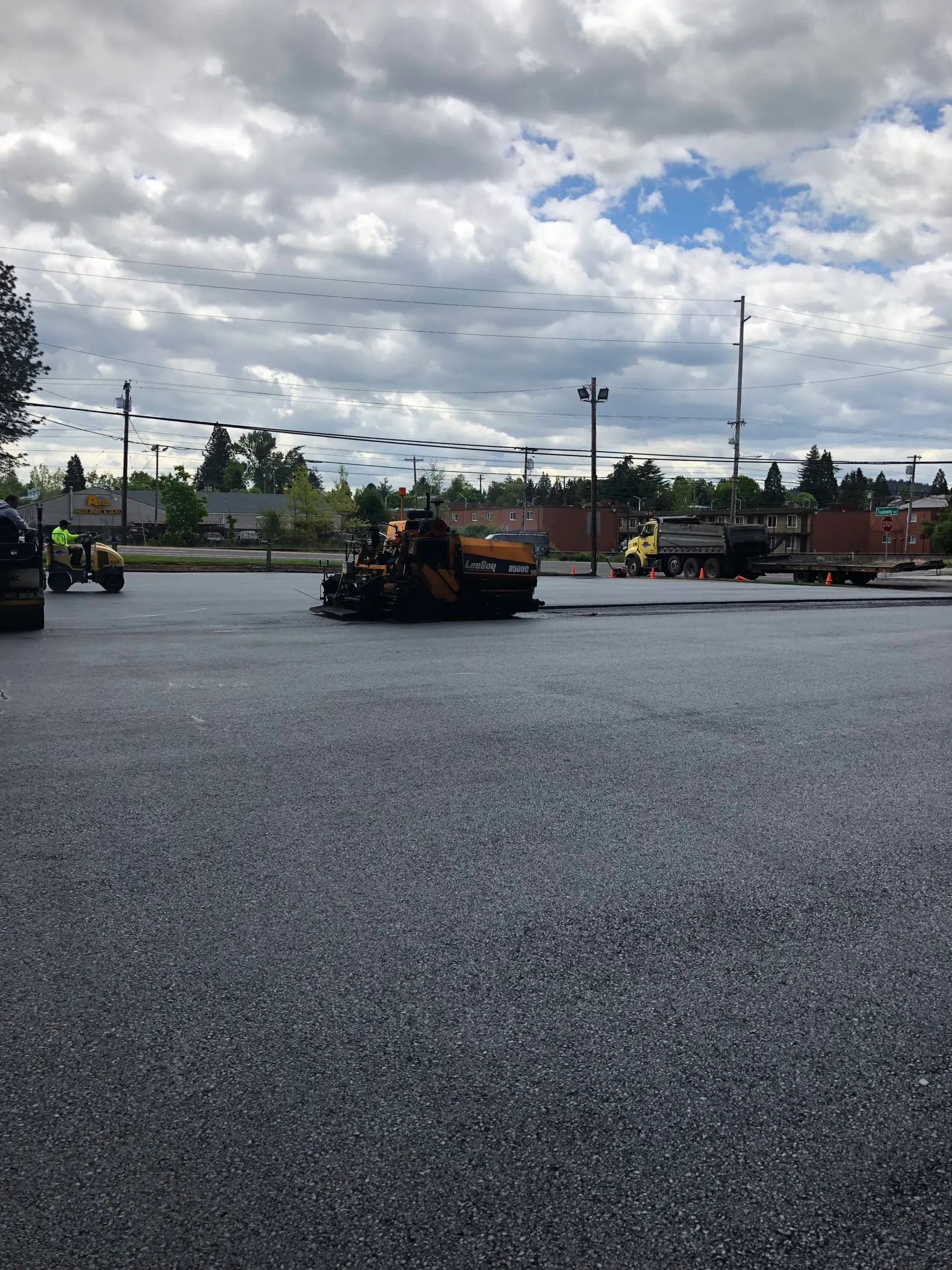 Construction equipment paves an asphalt parking lot under a cloudy sky.
