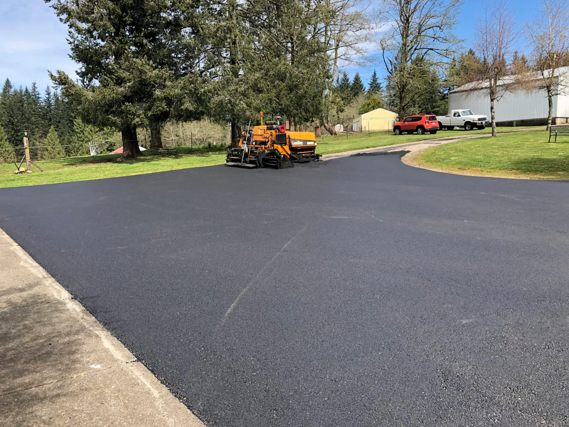 A freshly paved black asphalt driveway with a yellow construction vehicle parked near the trees.