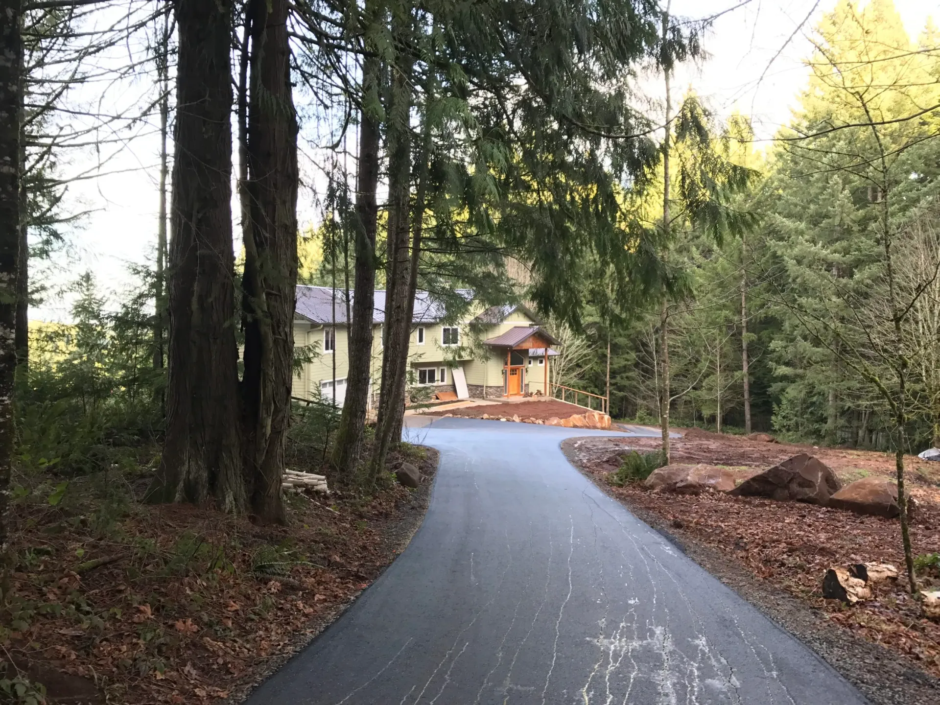 A paved driveway leads through a wooded area to a two-story house with a wooden entrance porch and surrounding forest.