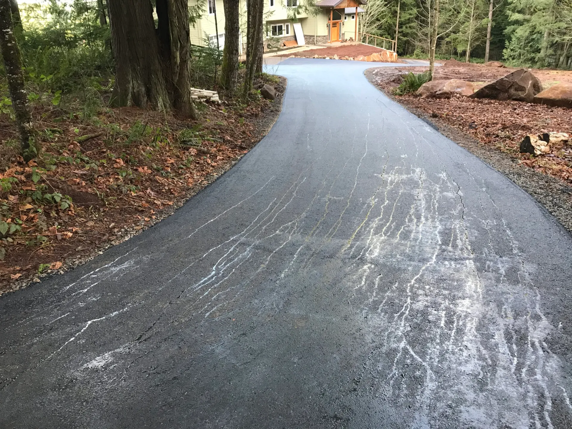 A freshly paved dark asphalt driveway curving through a wooded area toward a house in the distance.