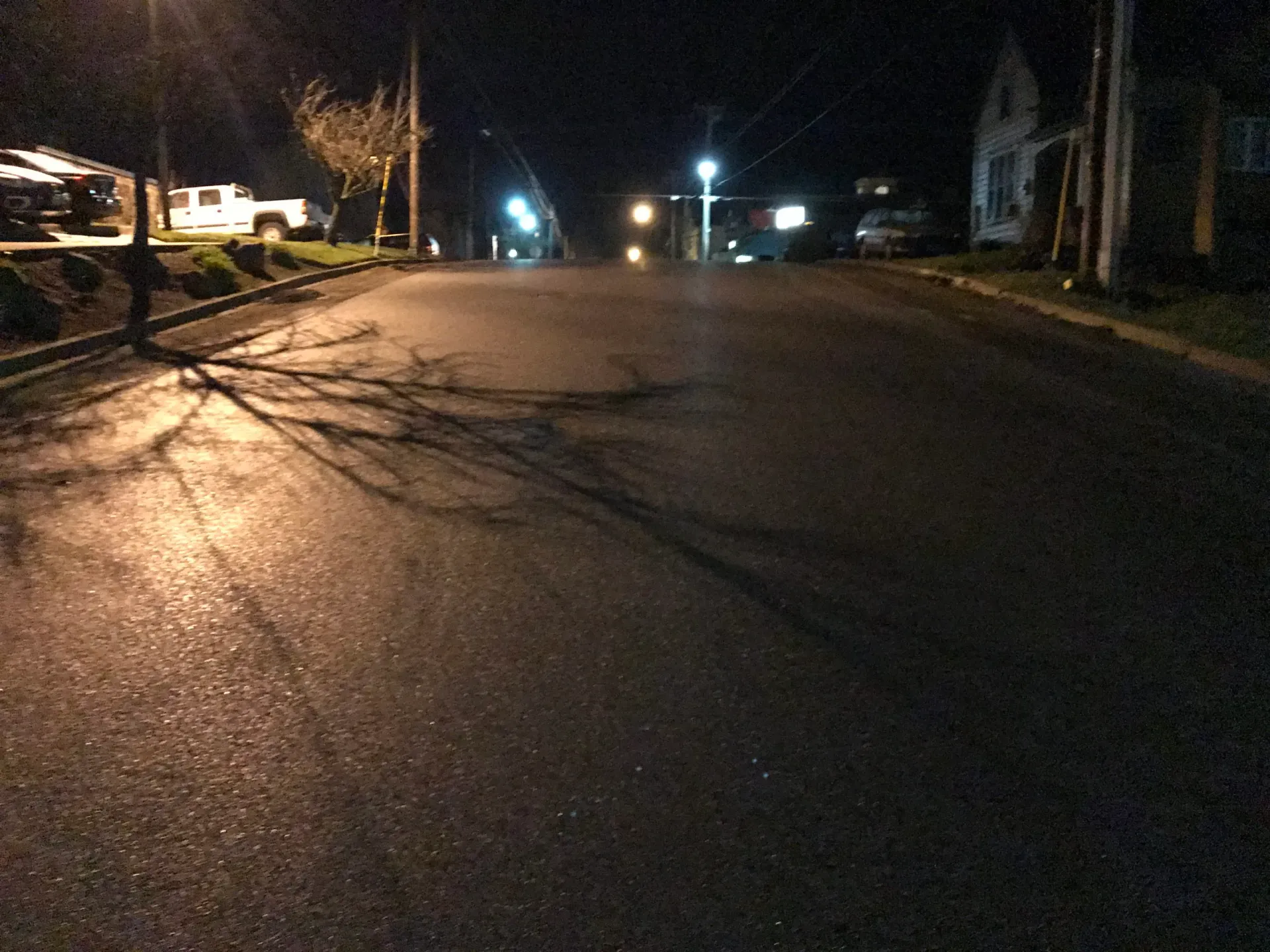 A night view of a dark, paved suburban street with glowing streetlights, tree shadows, and parked cars in the distance.