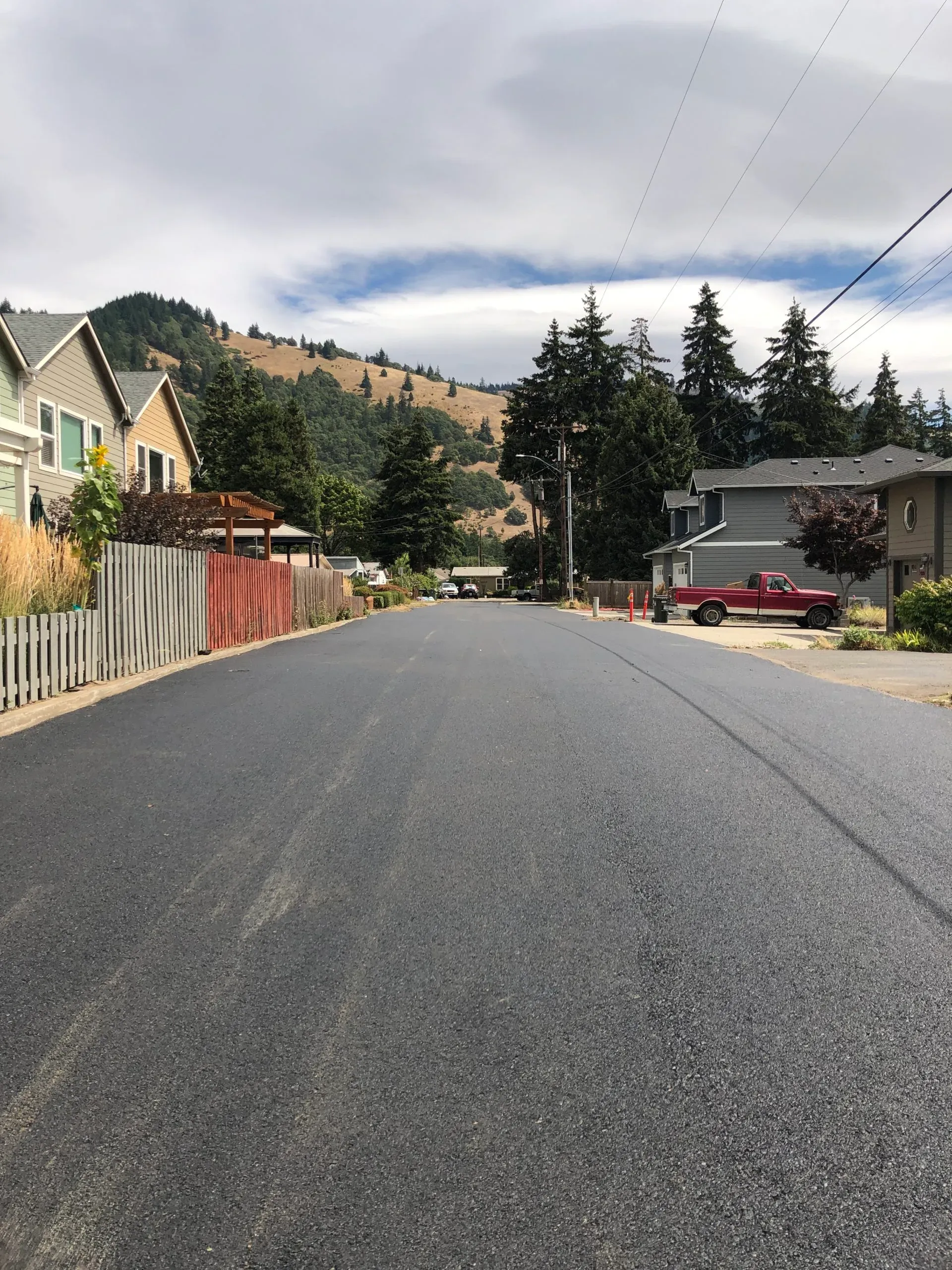 A quiet suburban street with freshly paved asphalt, residential houses, and a hilly forest backdrop under a cloudy sky.